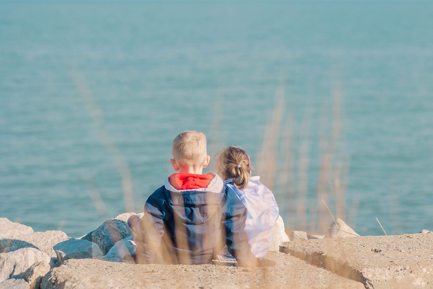 Two people sitting on coastal rocks overlooking calm blue waters on a bright day.