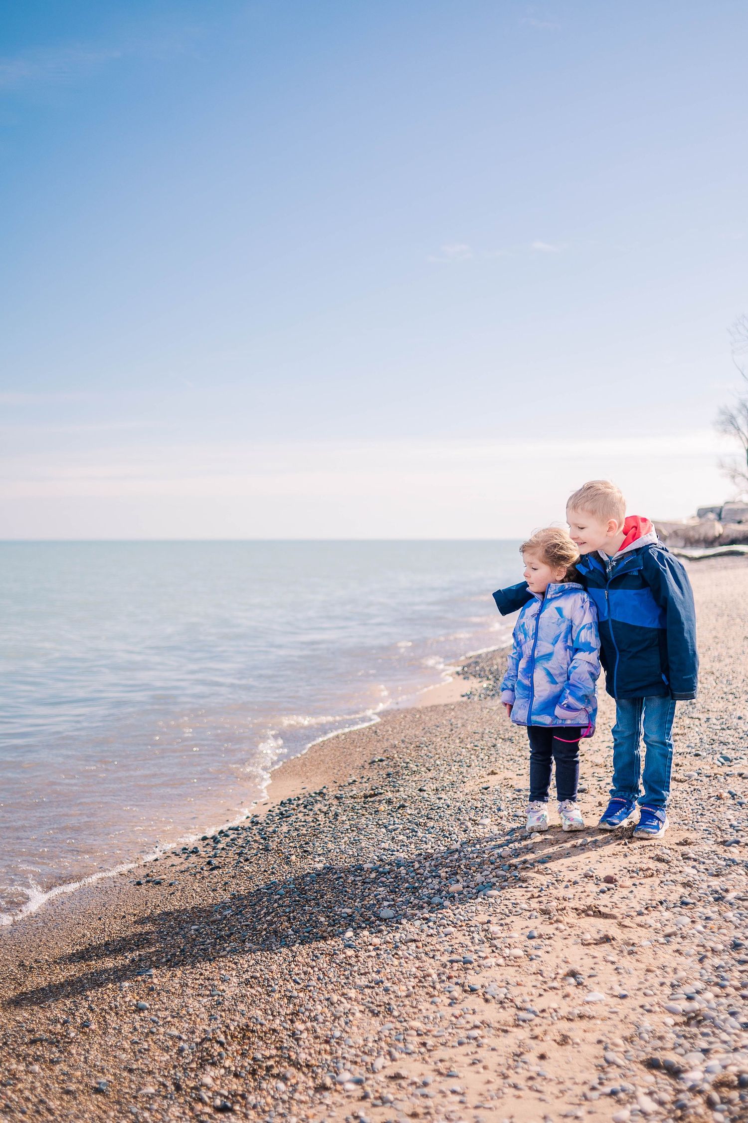 Two people in winter coats explore a rocky beach shoreline on a bright sunny day.