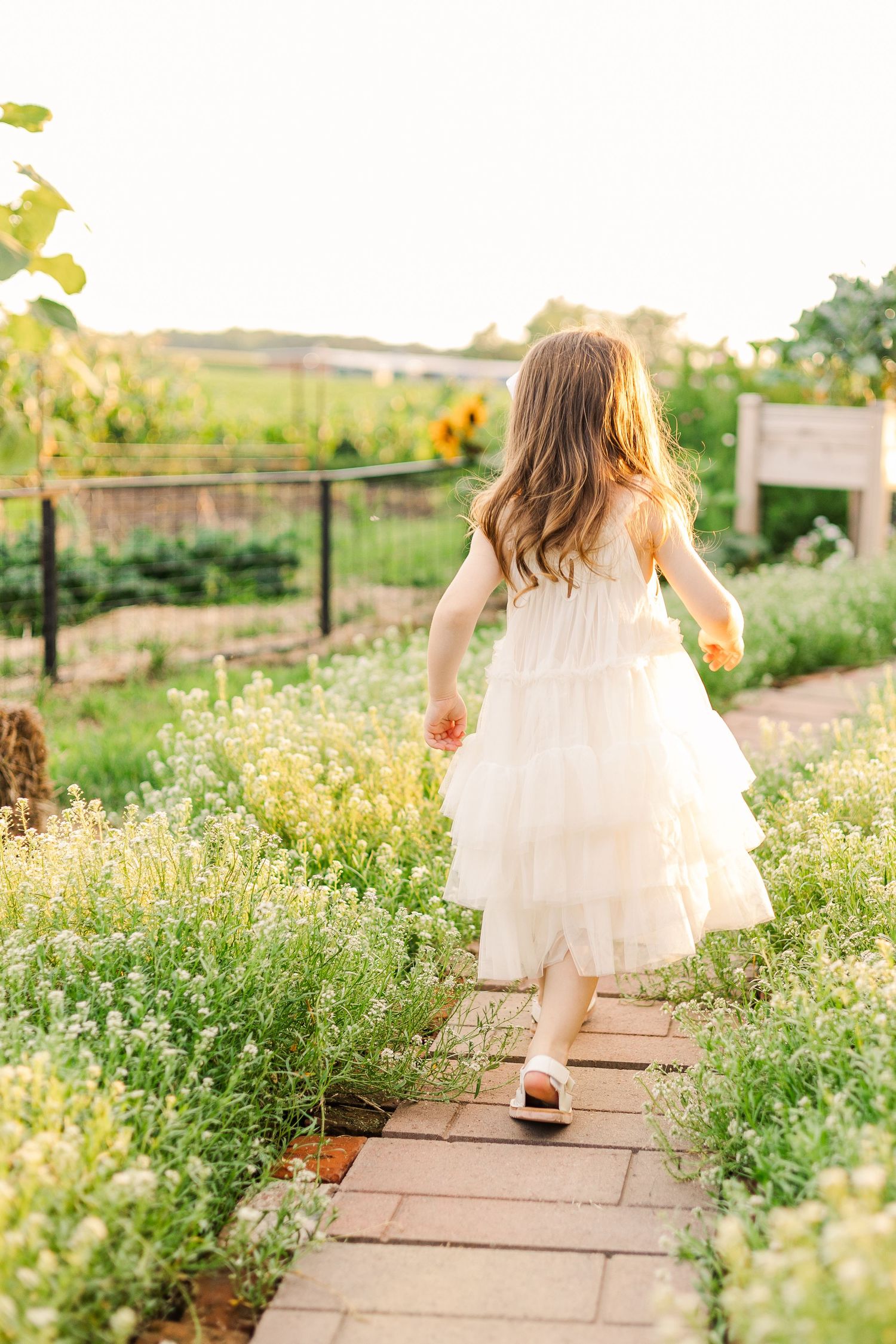 A child in a flowing white dress runs along a garden path surrounded by blooming flowers.