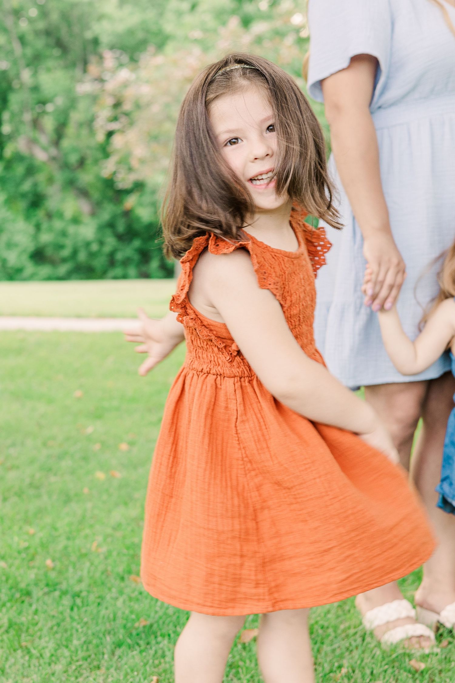 Young child in orange dress twirling playfully on grass during outdoor photoshoot.
