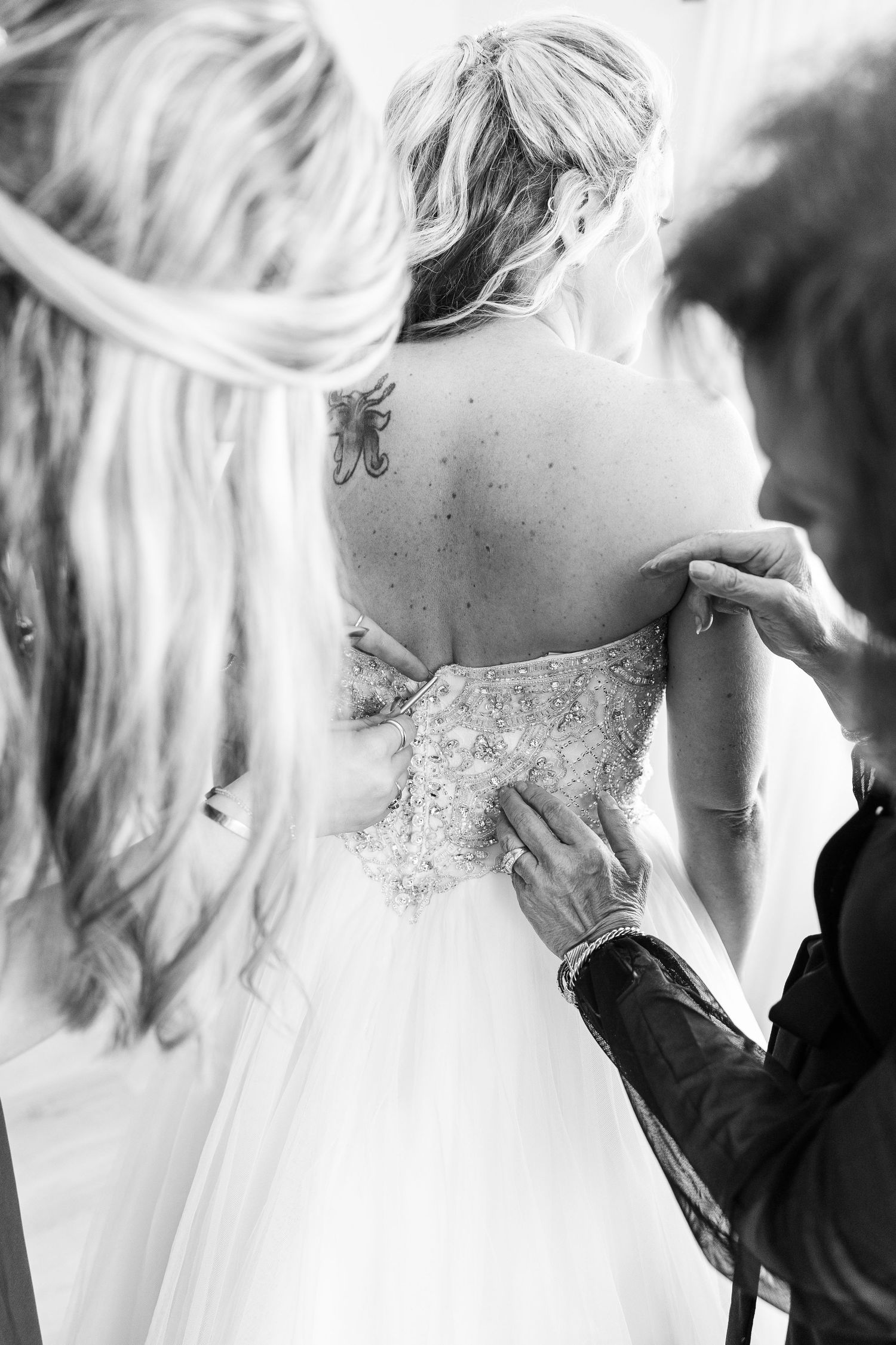 Black and white photo showing detail of ornate beaded wedding dress being laced up, with a small tattoo visible.