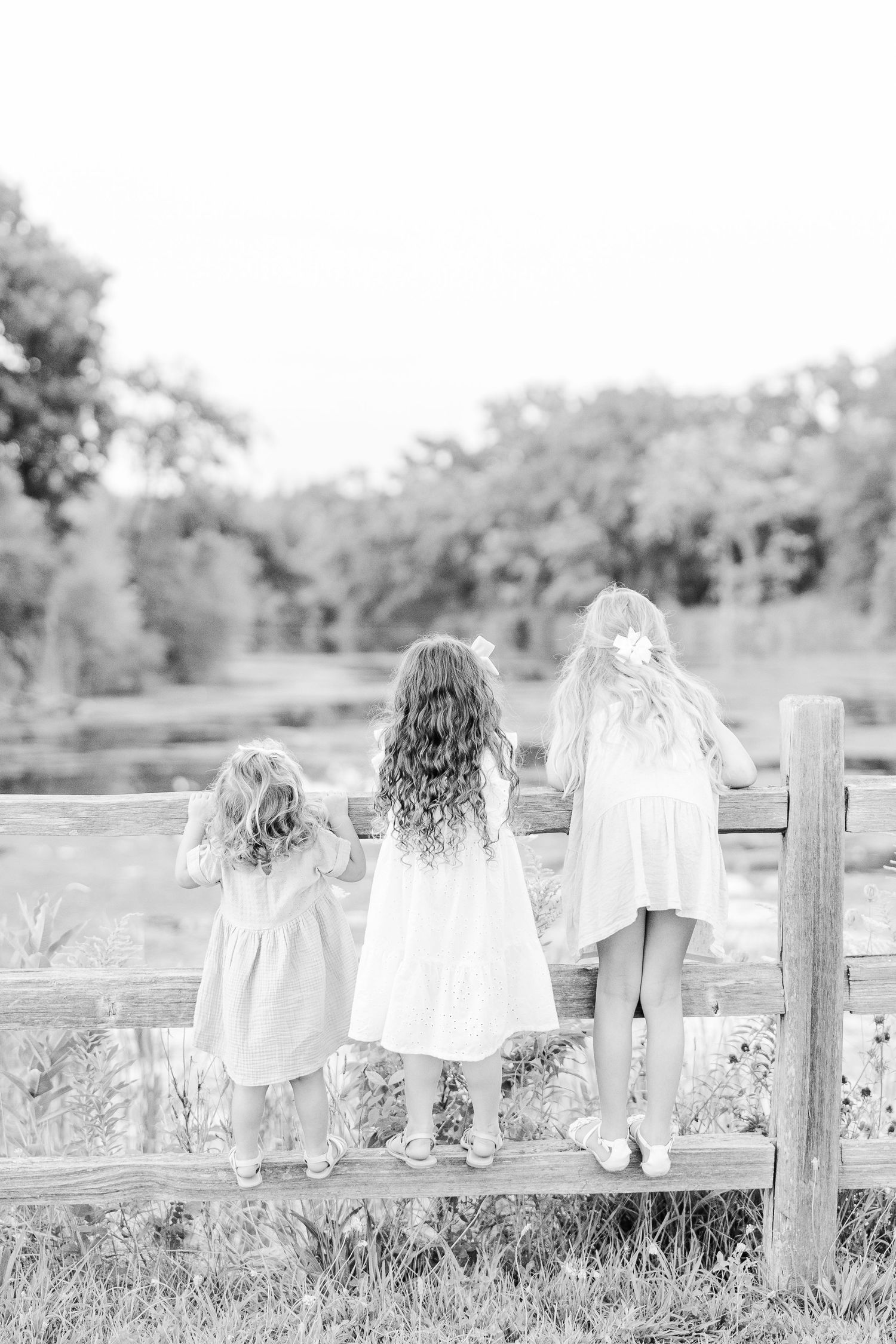 Three figures stand on wooden fence overlooking scenic pond in black and white photograph.