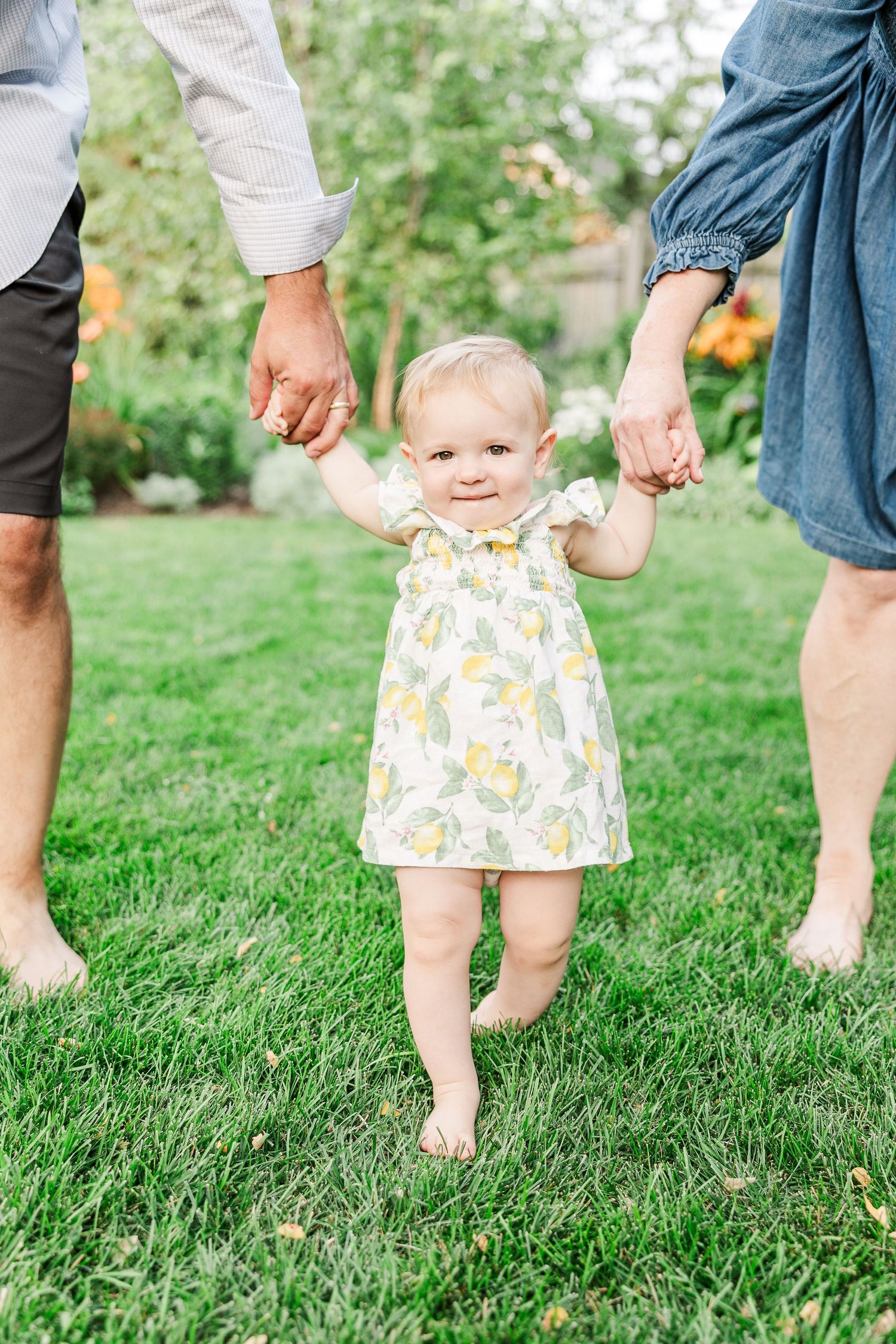 Small child in floral dress walking while holding hands with parents on green lawn.