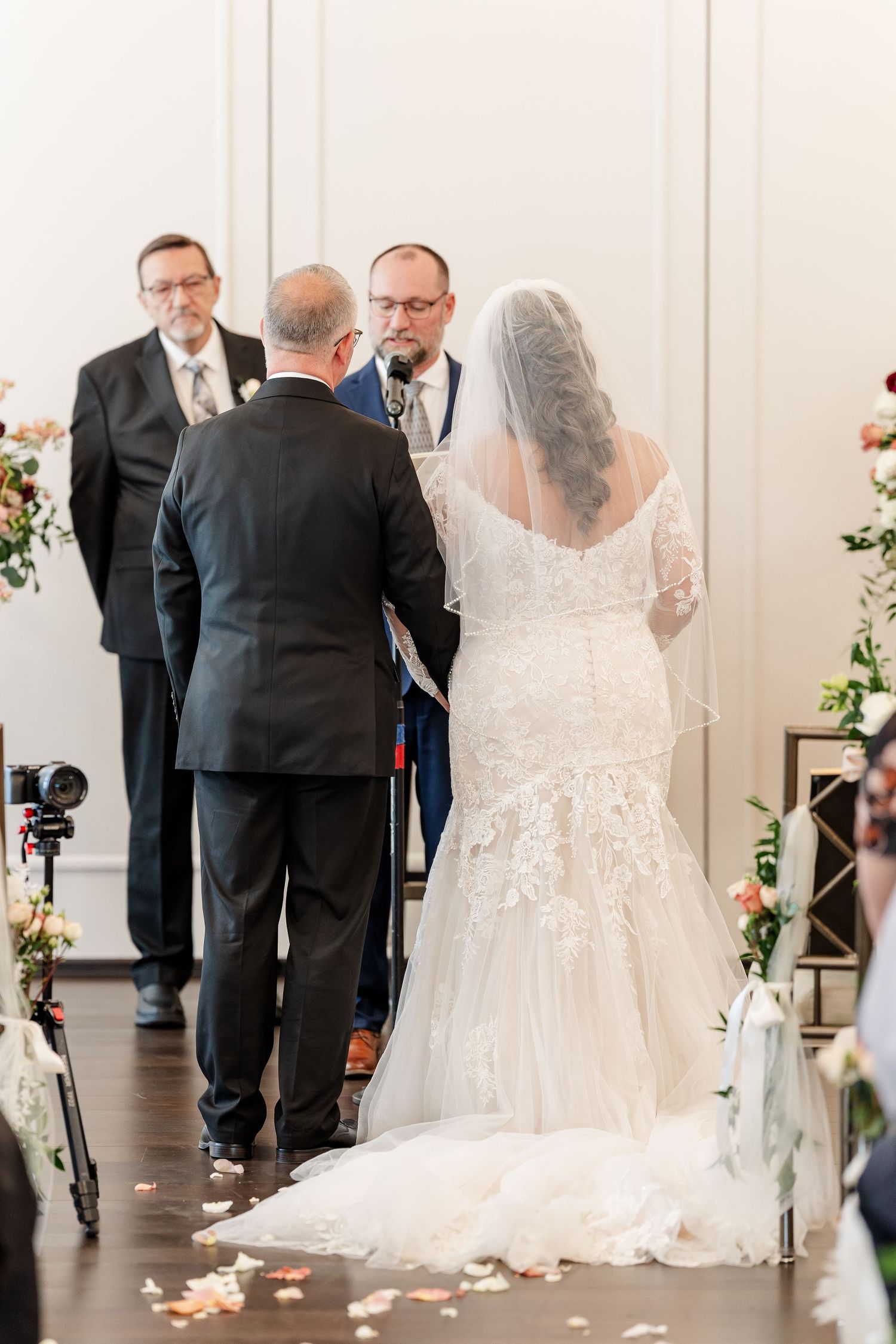 Bride in a lace wedding gown and long veil faces the groom as an officiant stands at a microphone during the ceremony.