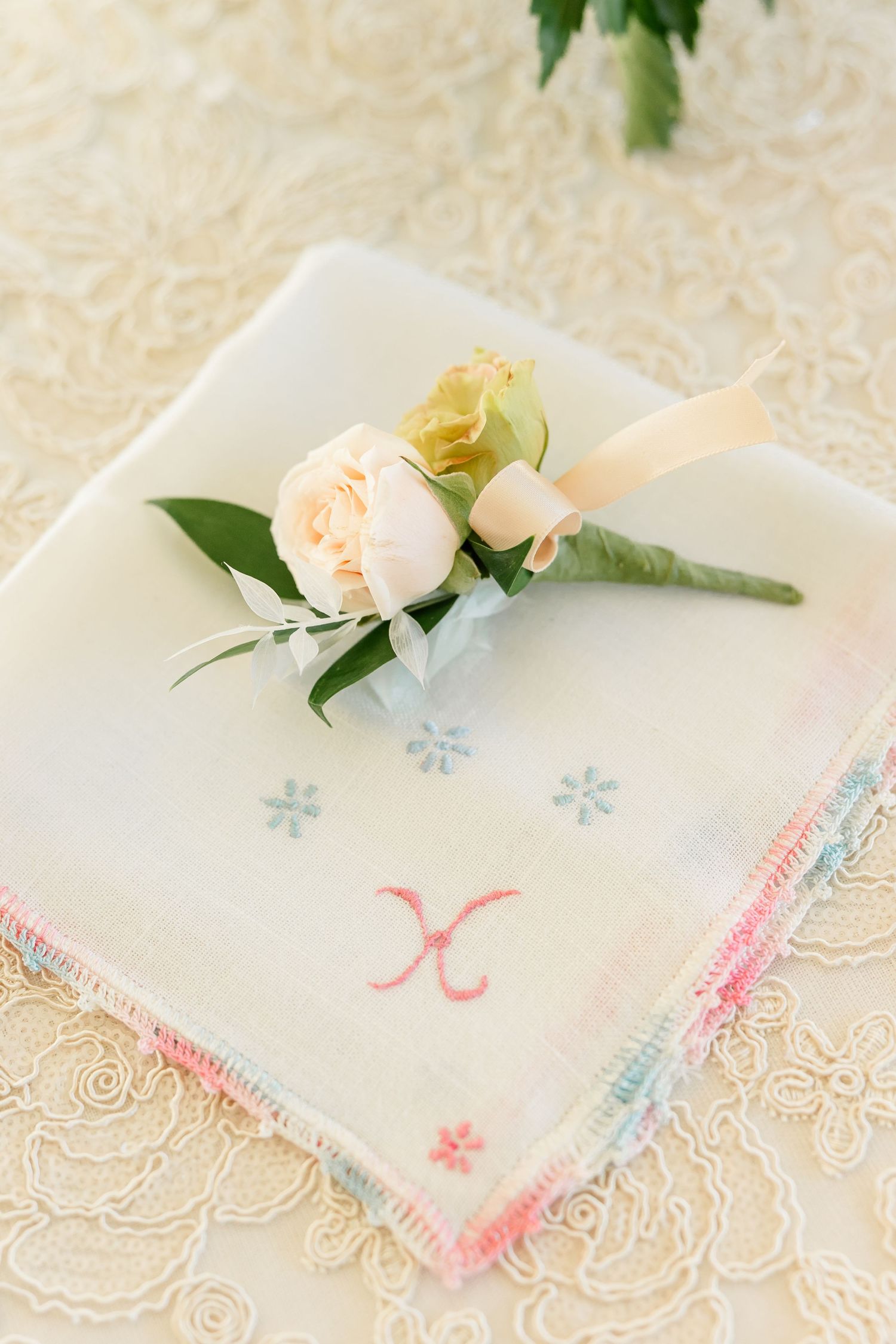 Peach rose boutonniere with a ribbon on a vintage embroidered handkerchief with pastel edging, resting on ivory lace.