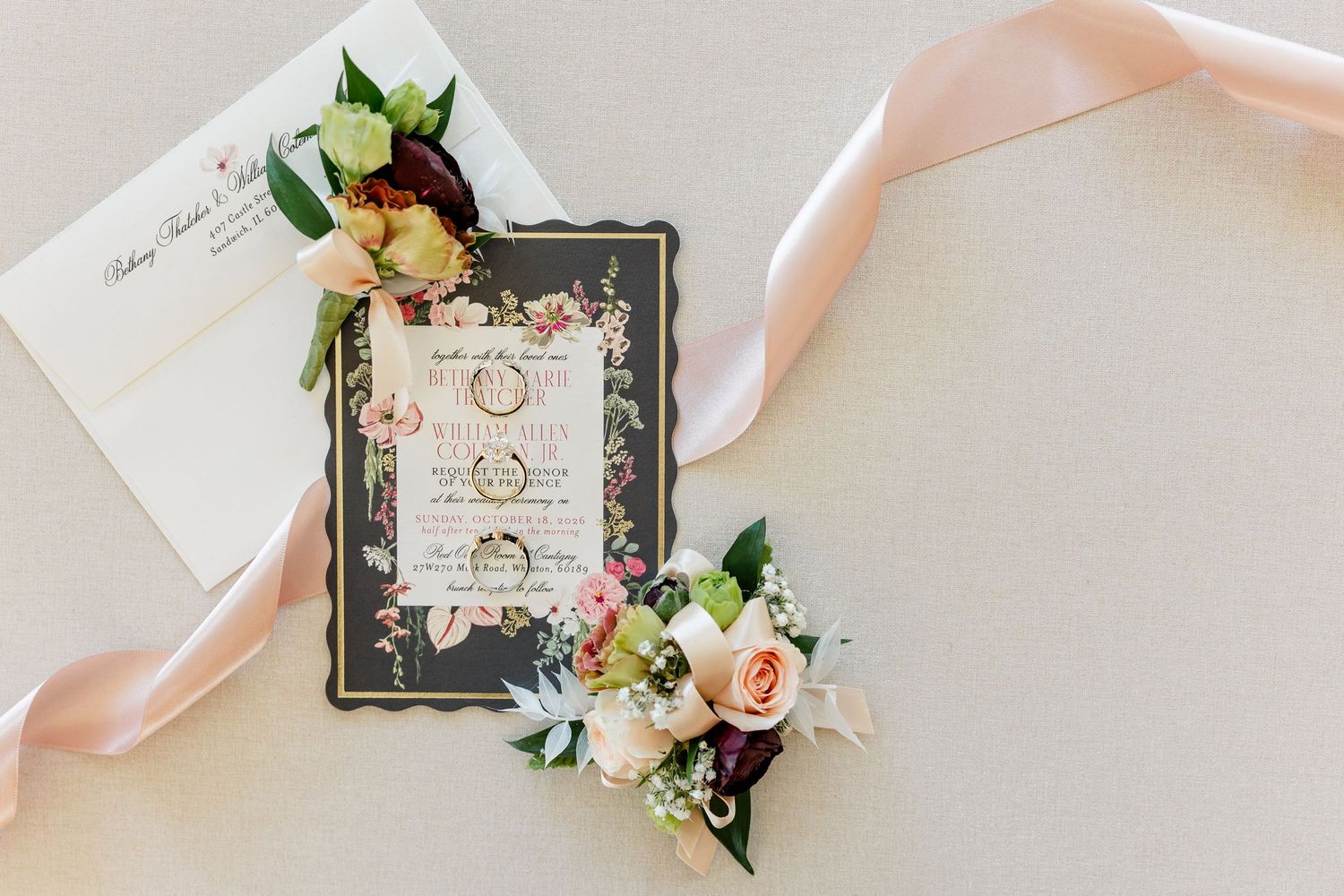 Black and gold floral wedding invitation with two rings, surrounded by blush ribbon, a peach rose boutonniere, and a pink floral corsage.