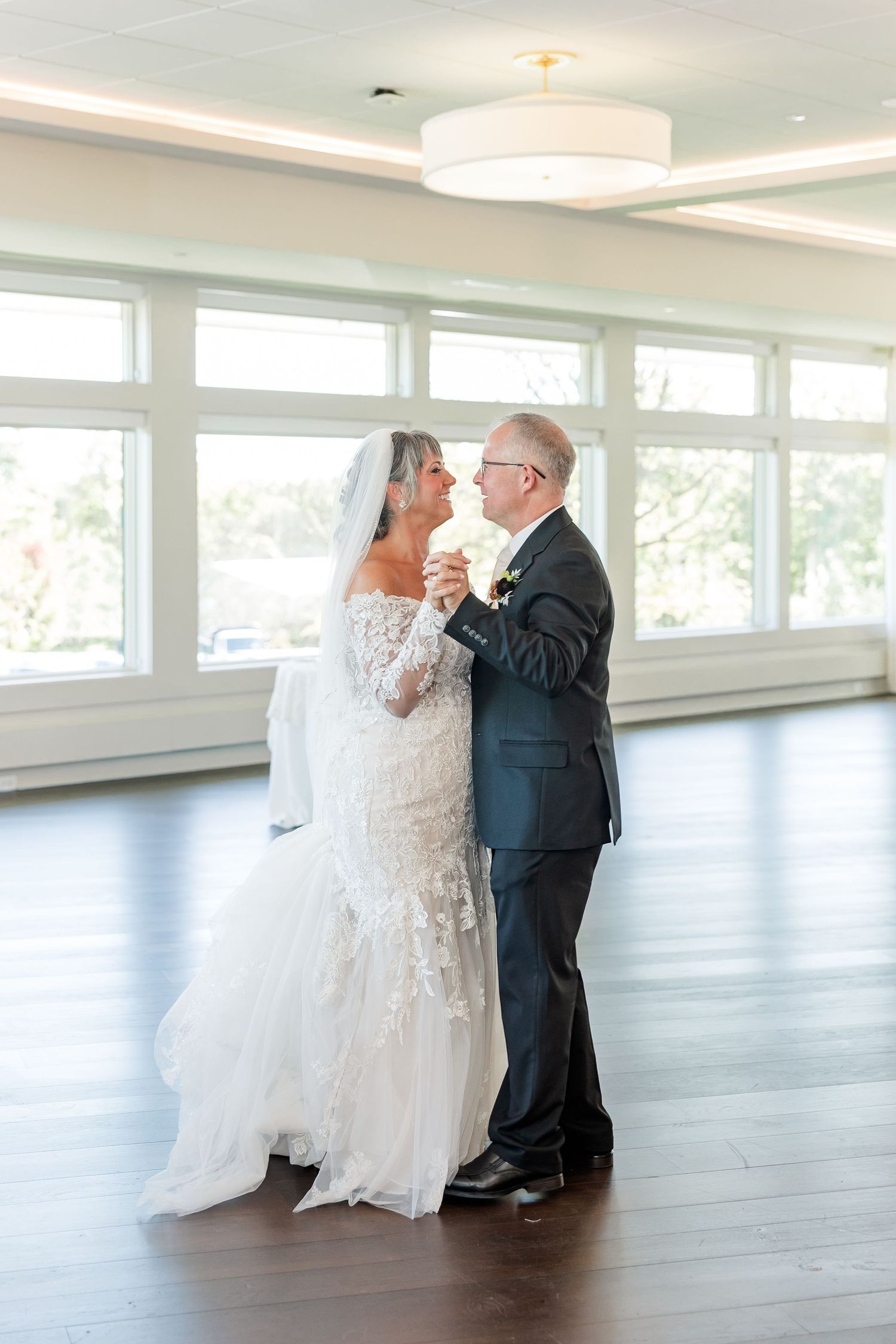 A bride in a white lace gown and veil shares a first dance with her groom in a navy suit in a bright ballroom with large windows.
