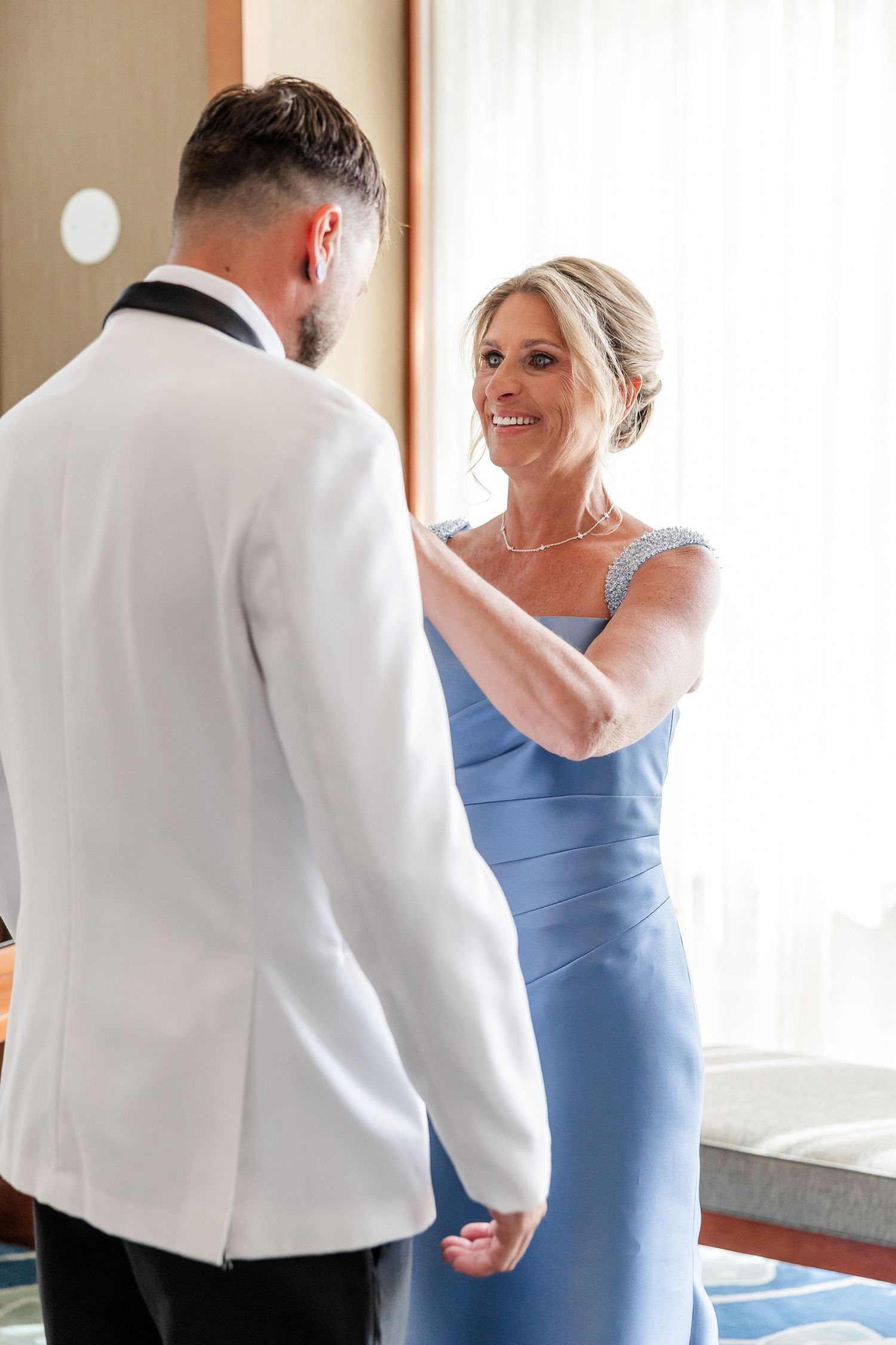 A woman in a blue gown adjusts the lapel of a groom in a white tuxedo jacket before the wedding ceremony.