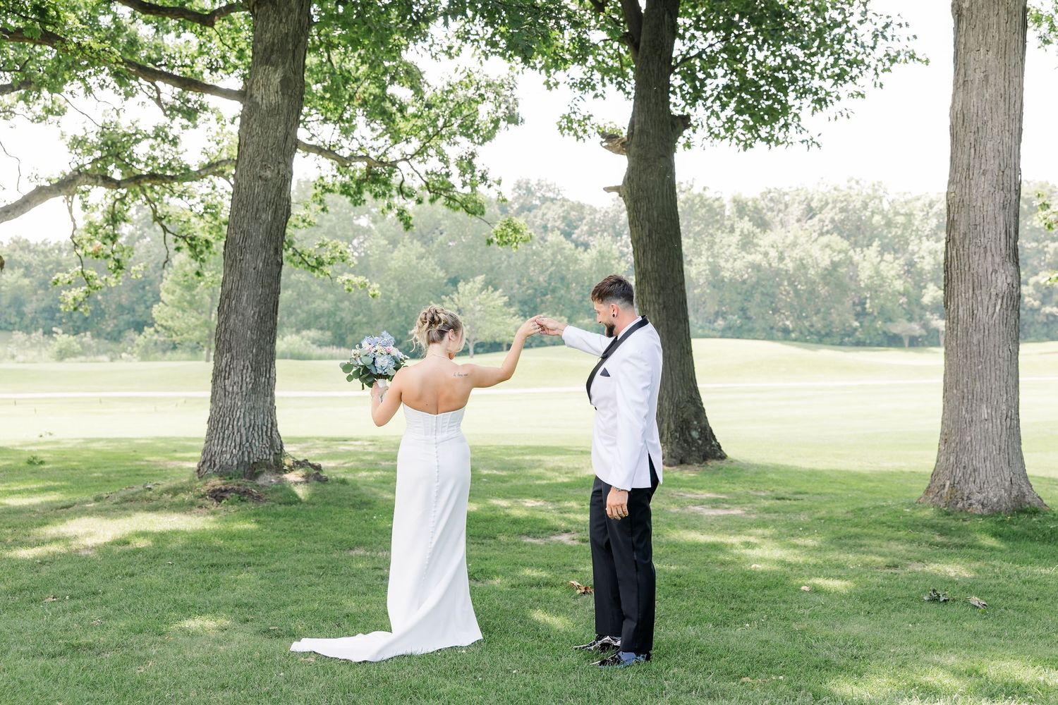 Bride and groom share a romantic first look moment under trees on a sunny green lawn at outdoor wedding.