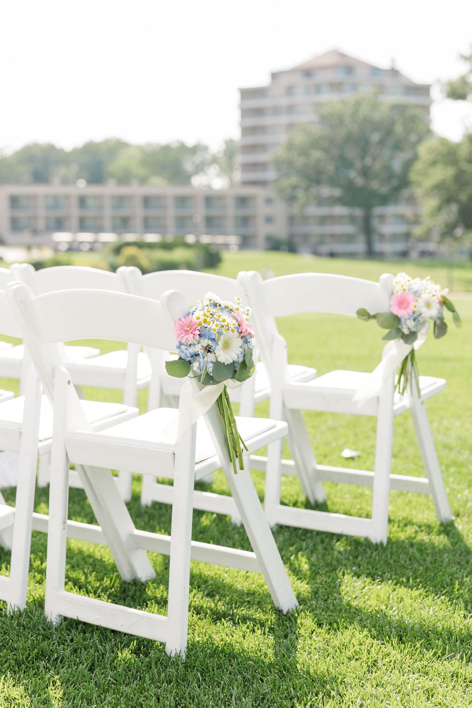 White folding chairs decorated with colorful floral bouquets line the aisle of an elegant outdoor wedding ceremony.