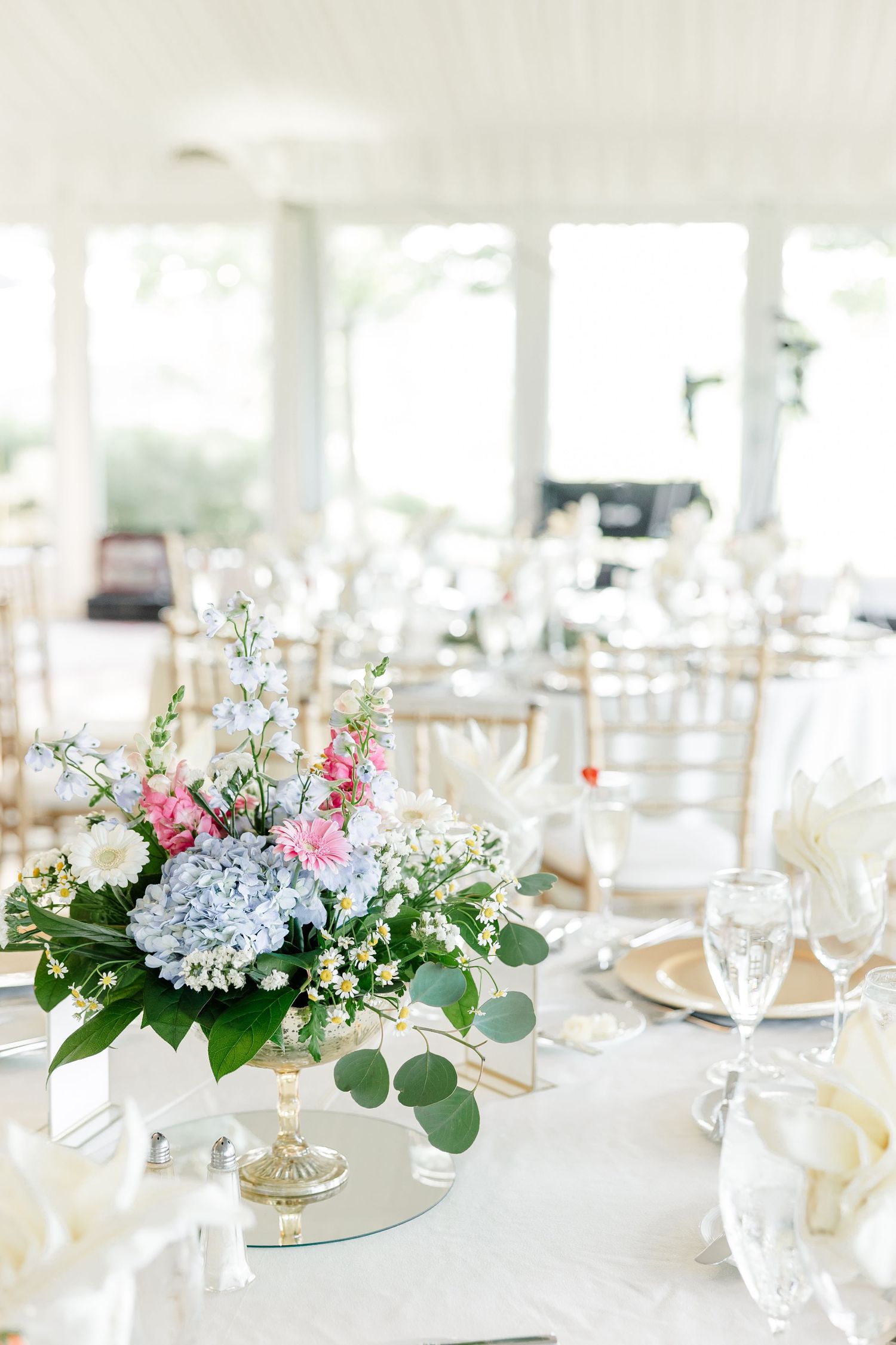 Elegant wedding reception table with a floral centerpiece of blue hydrangeas and pink flowers in a bright venue.