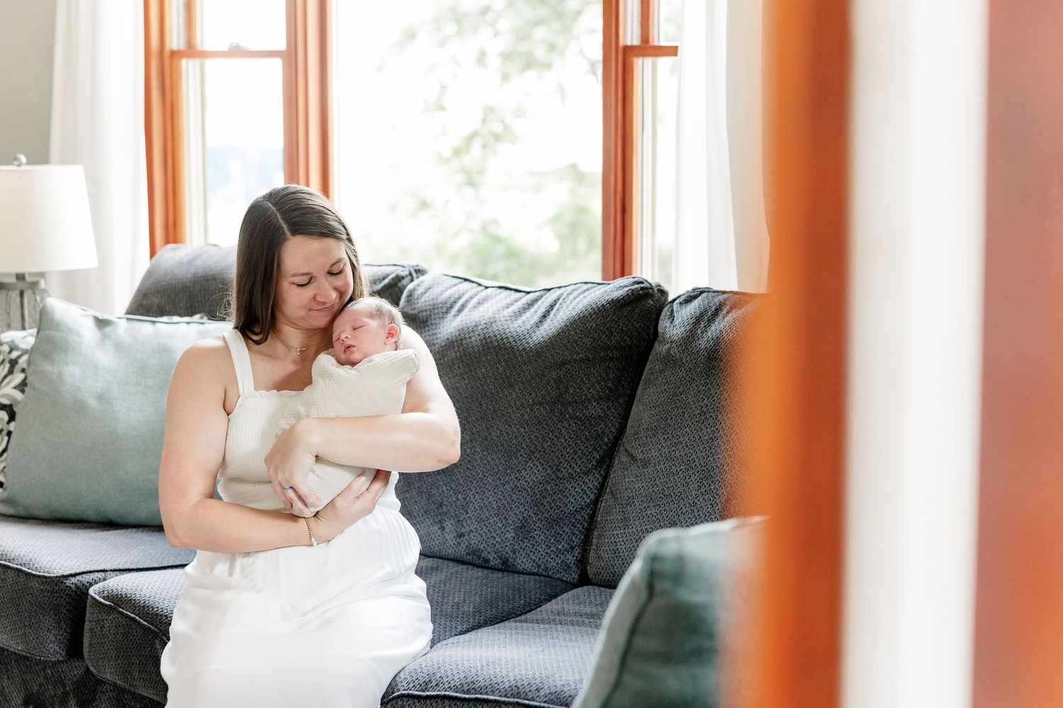 A mother in a white dress tenderly holds her newborn baby while sitting on a gray sofa in a bright living room.