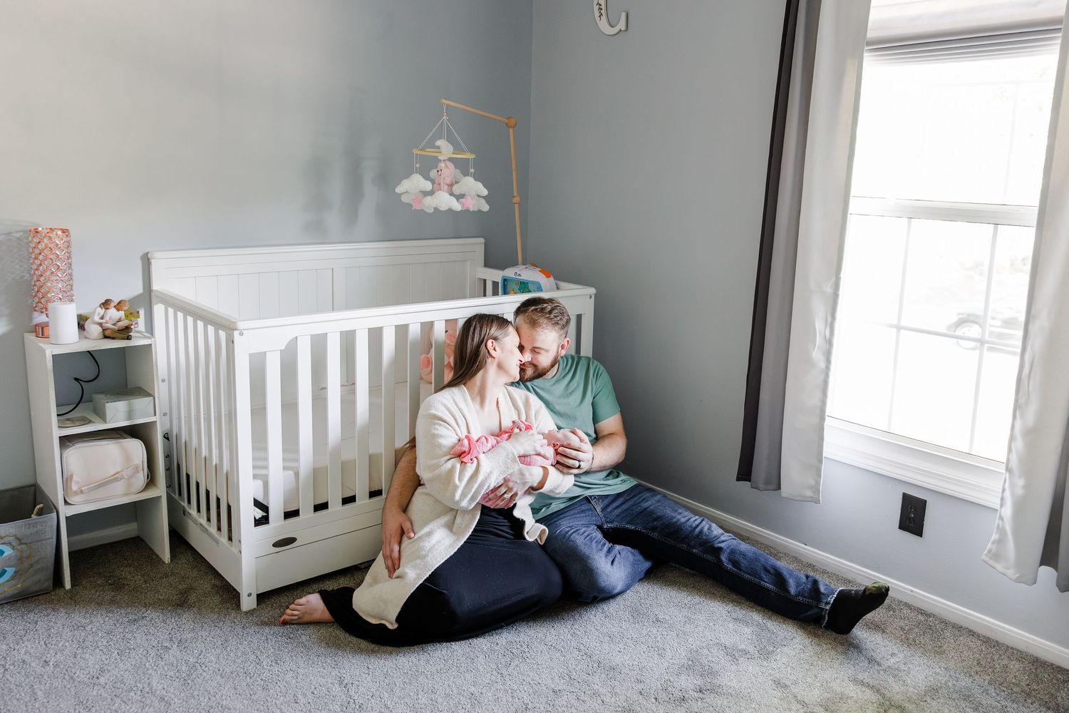 New parents sit on a nursery floor holding their newborn baby next to a white crib in a gray room.