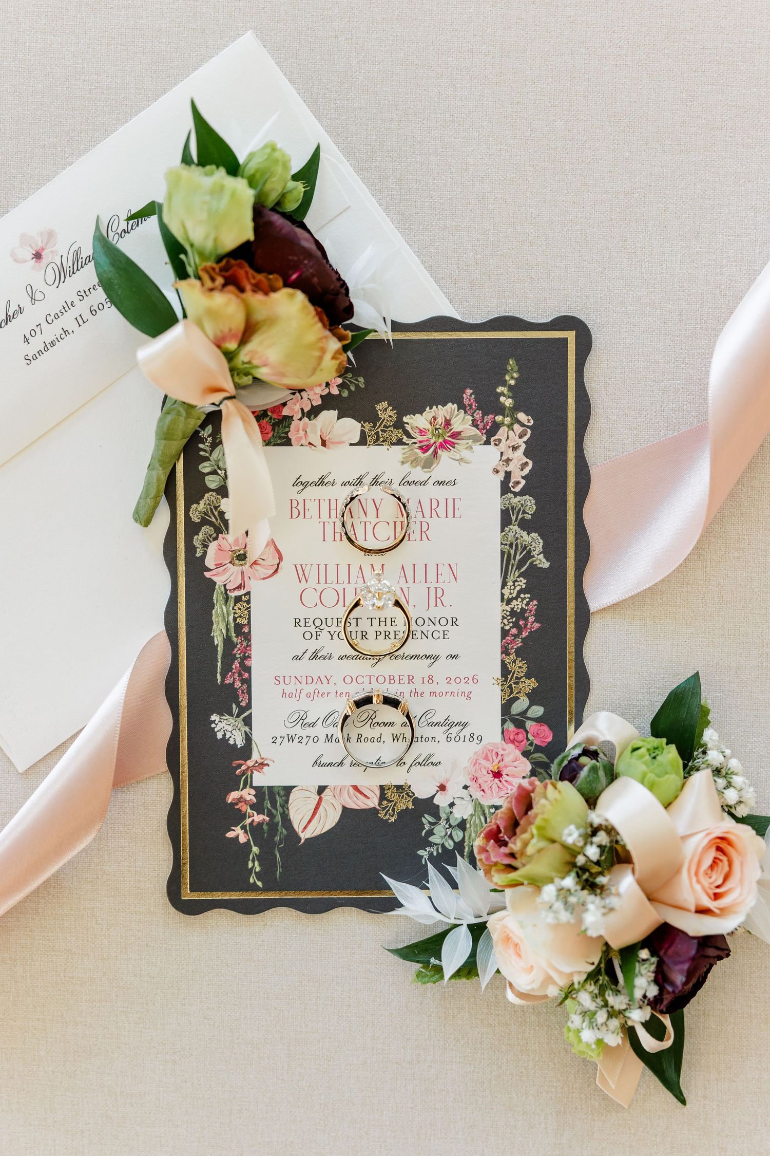 Elegant black and gold floral wedding invitation with blush ribbon, envelope and pink rose boutonnieres on a neutral background.