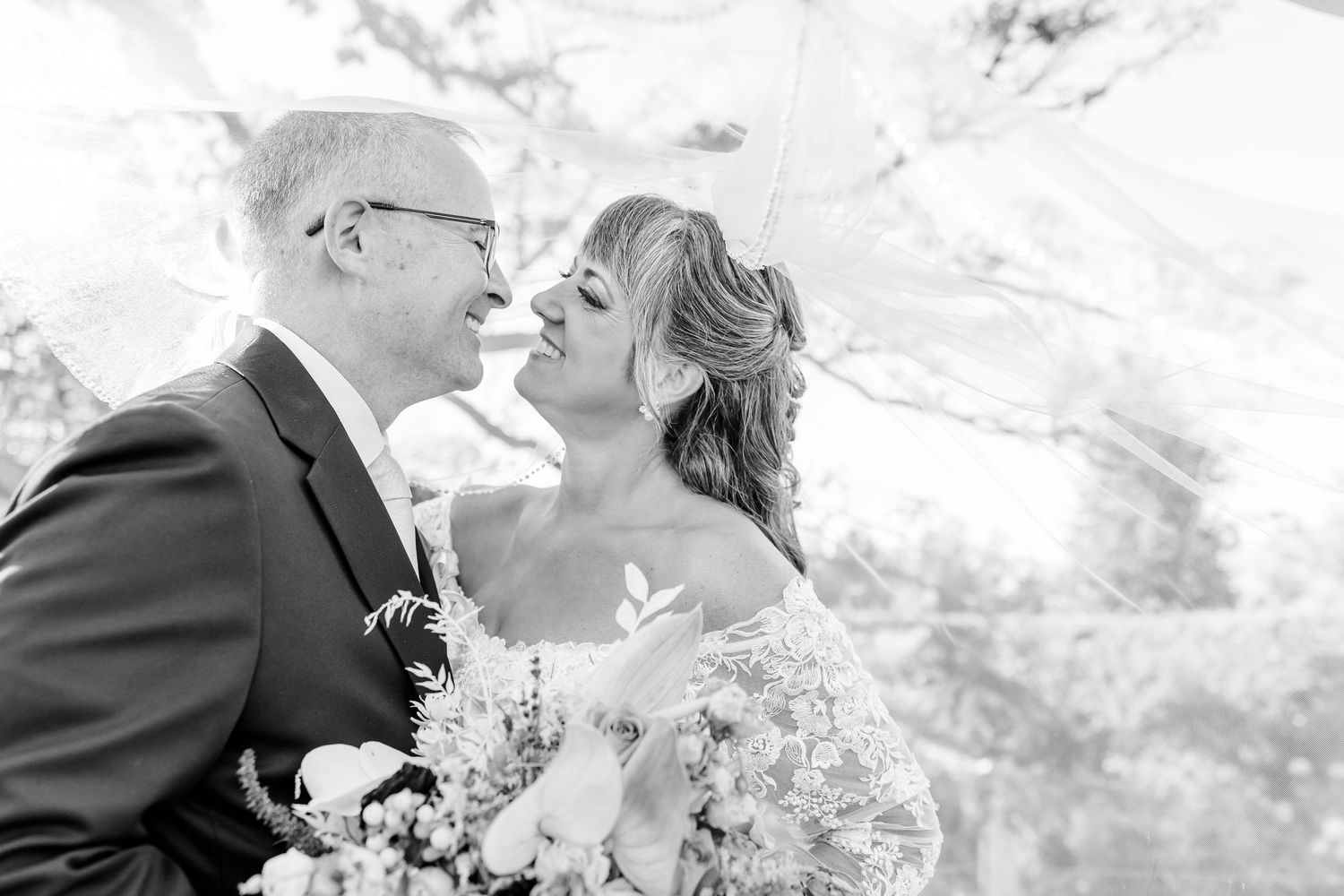 Black and white photo of a smiling bride and groom sharing a tender moment under a sheer veil, with the bride holding a bouquet.