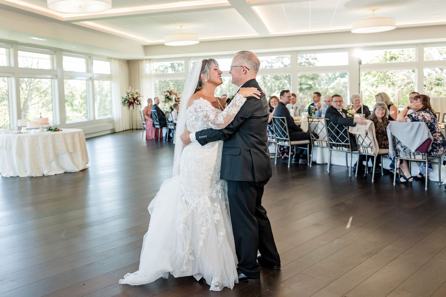 A bride in a white lace gown and groom in a dark suit share a romantic first dance on a wood floor while wedding guests watch.