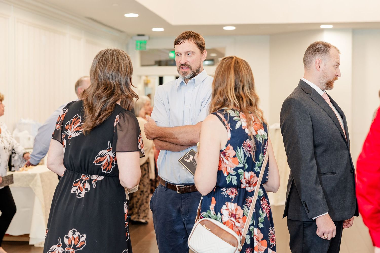 Wedding guests in floral dresses and suits chat and mingle together in a bright, elegant reception venue.