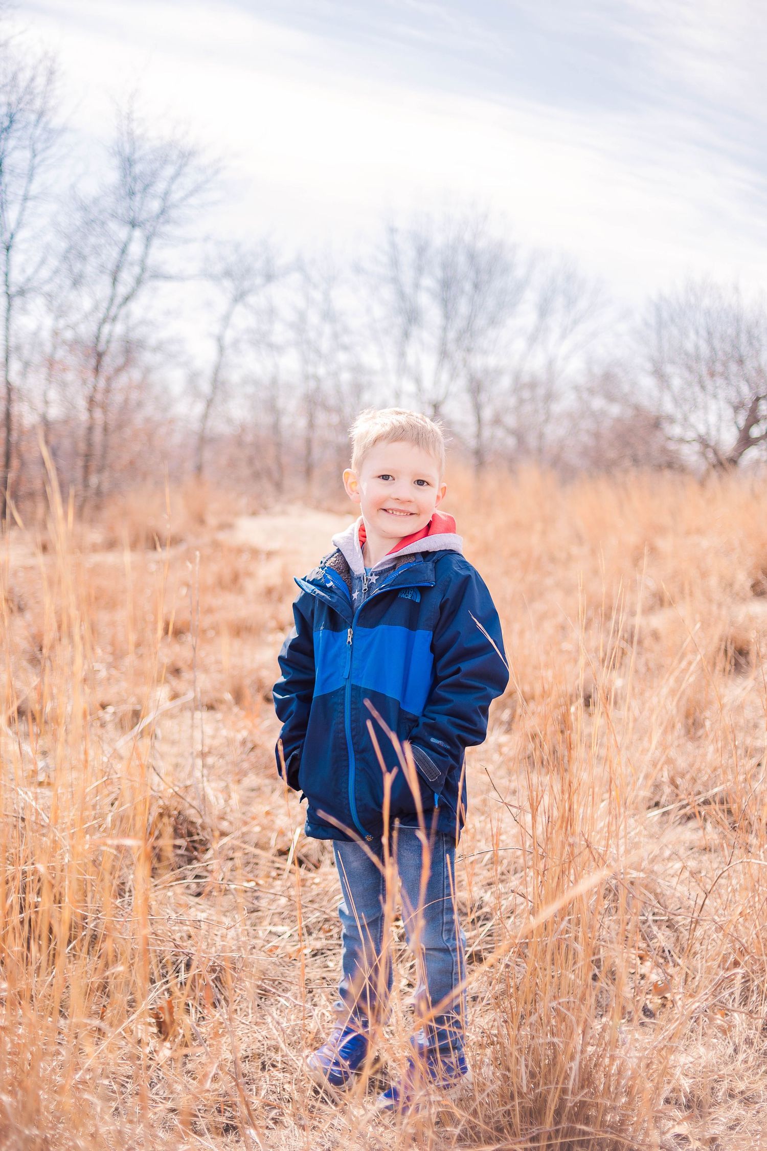 A young person stands in tall grass wearing a blue winter jacket on a cold autumn day.