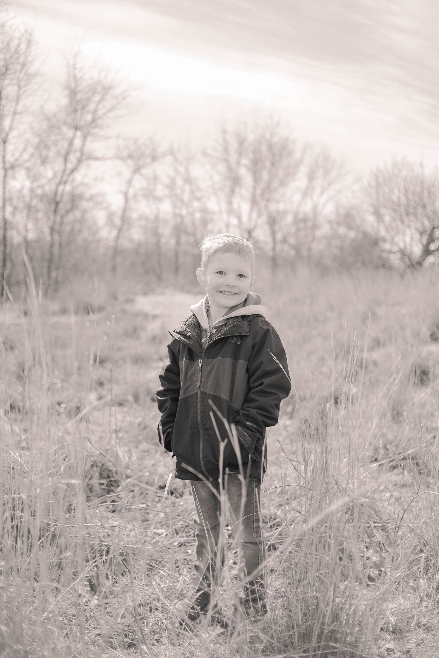 A black and white portrait of someone standing in a field of dried grass and bare trees.