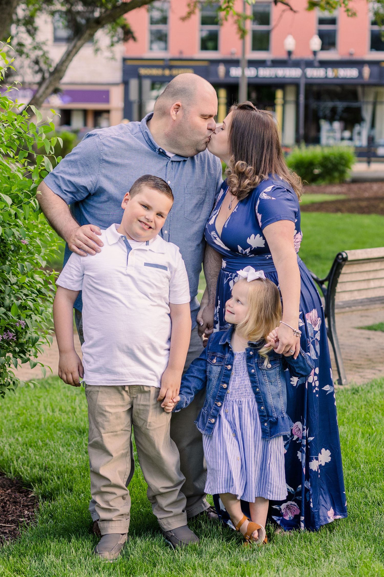 Family of four poses together on grass with pink building and trees in background.