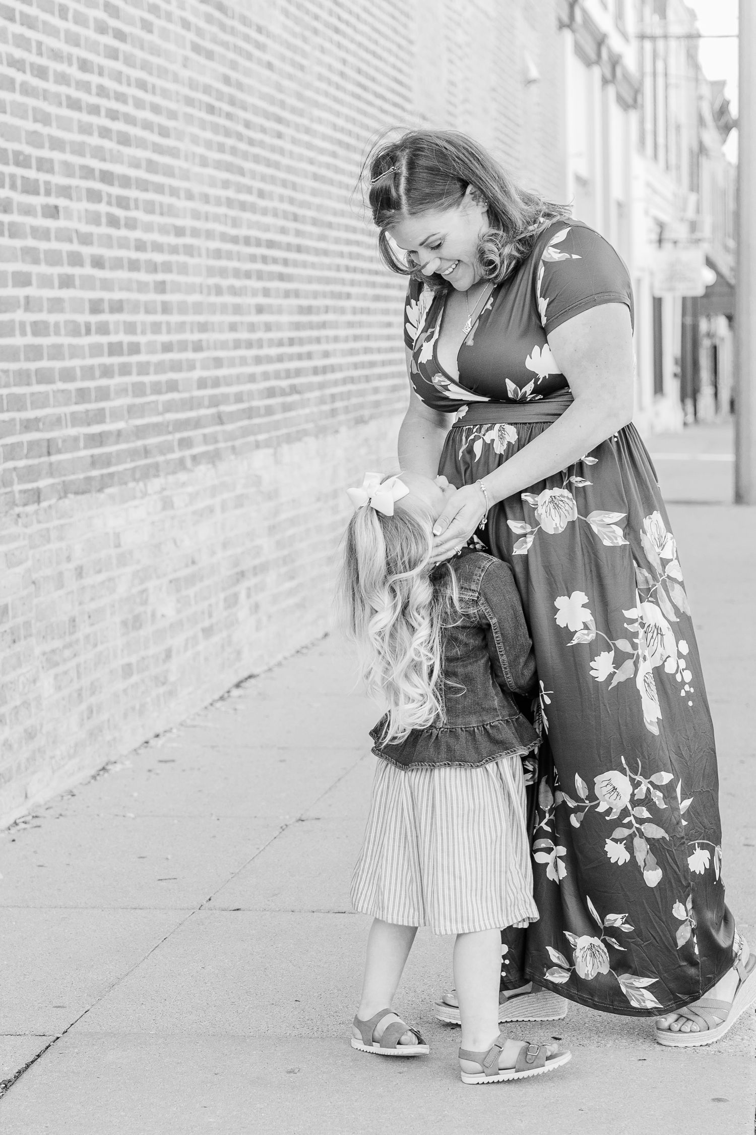 Woman in a floral dress bends down to interact with a child in a striped outfit against brick wall.