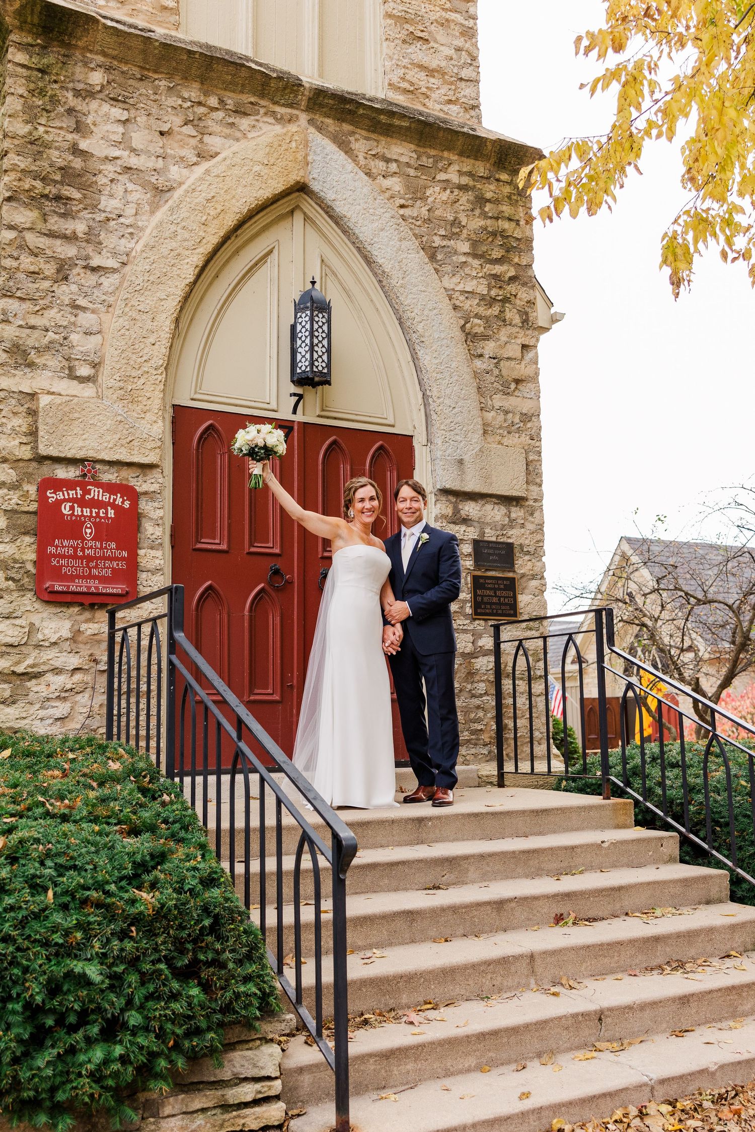 Wedding couple poses on stone church steps in front of a red wooden door with Gothic archway.
