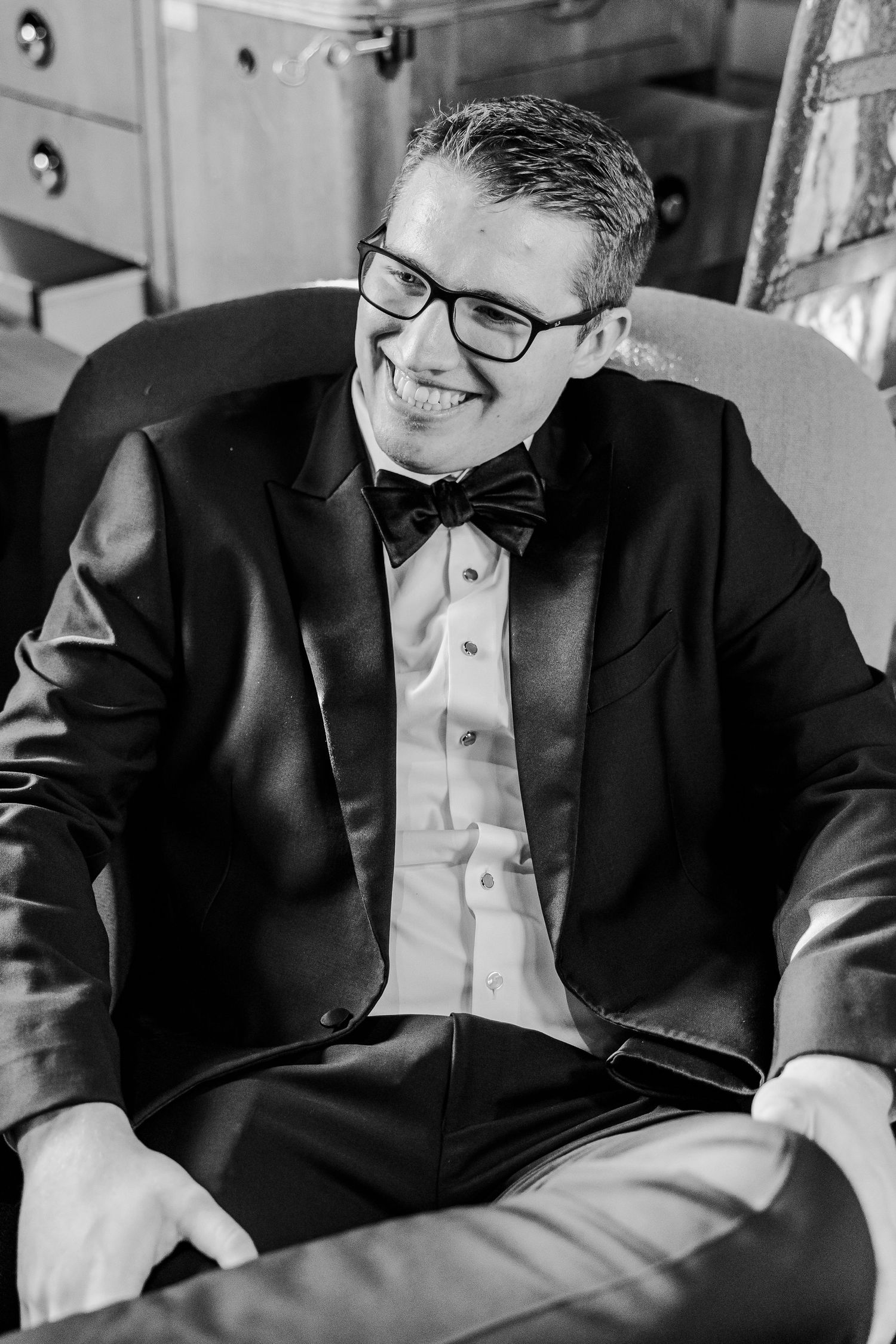 A smiling groomsman in glasses and tuxedo sits casually in a chair.