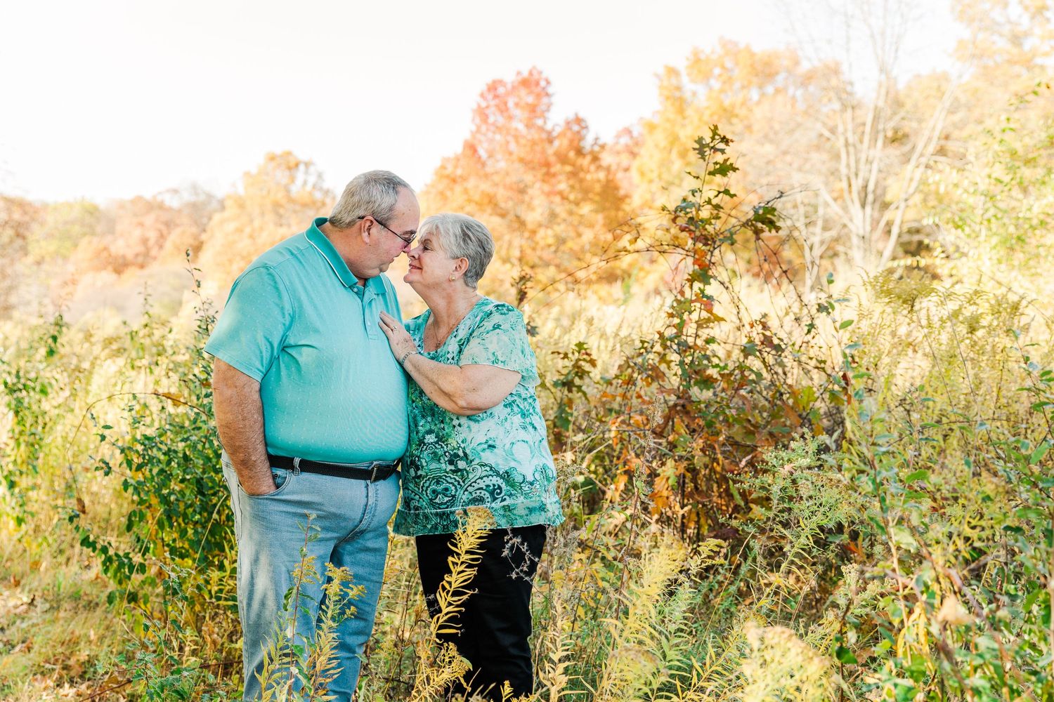 An elderly couple shares a tender moment in a field during golden hour with autumn colors.