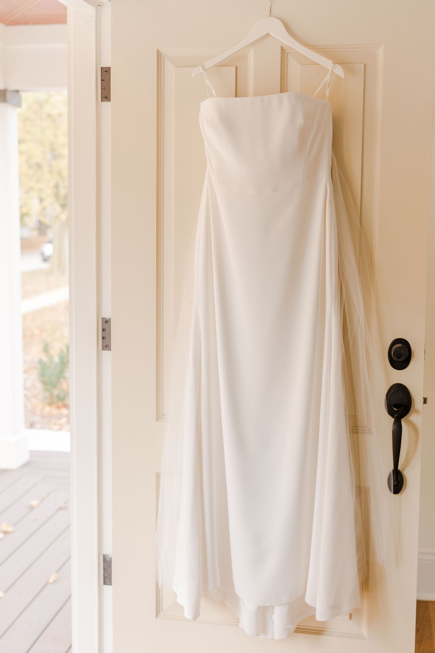 Sleek white wedding dress hanging on a white door in natural light.