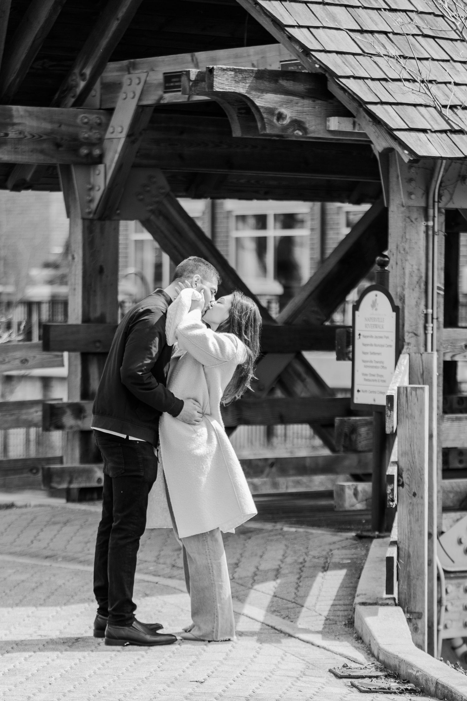 A romantic black and white photo of a couple embracing under a rustic wooden covered bridge structure.