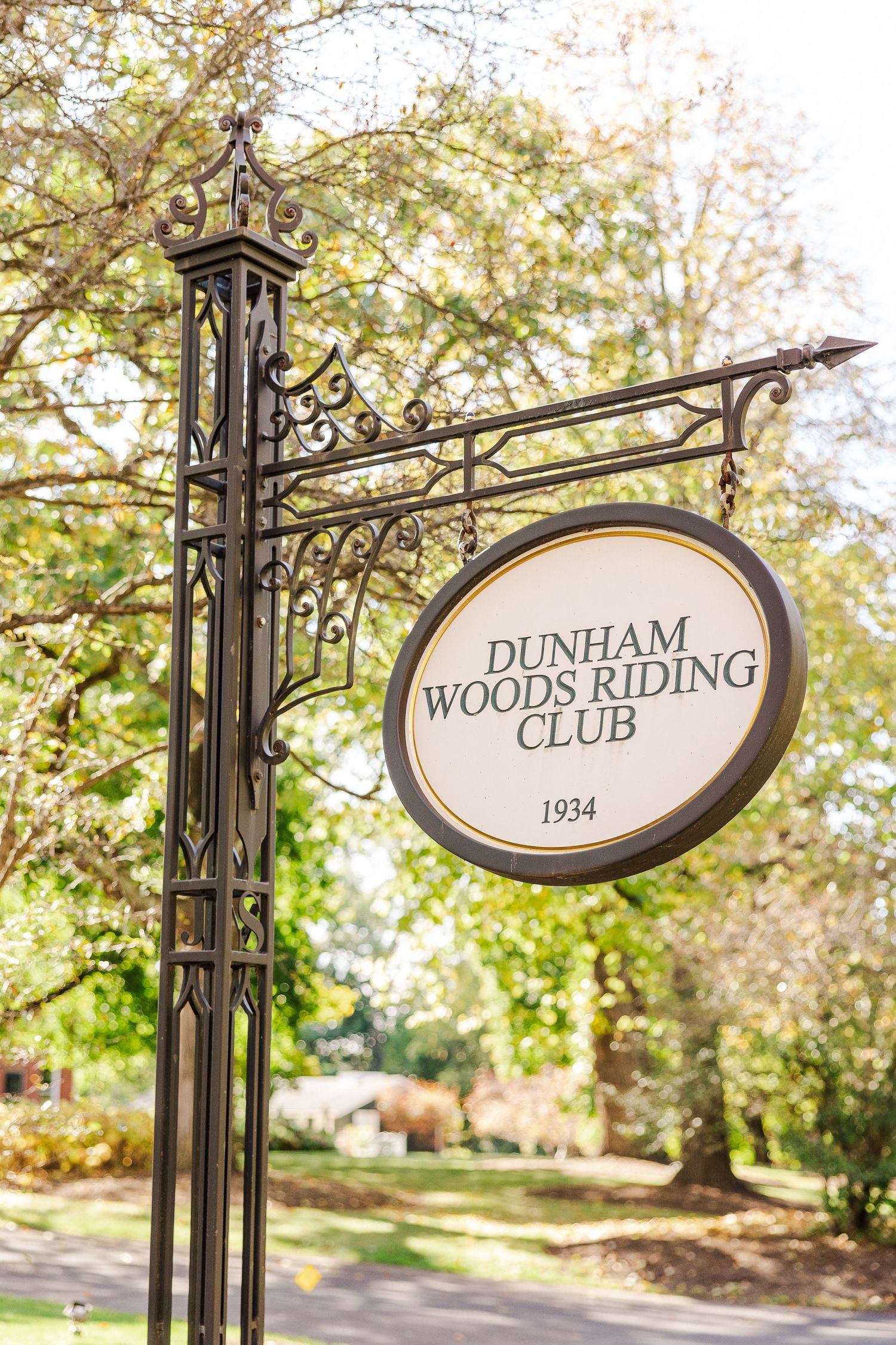 Ornate wrought iron sign for Dunham Woods Riding Club established in 1934 against autumn foliage.