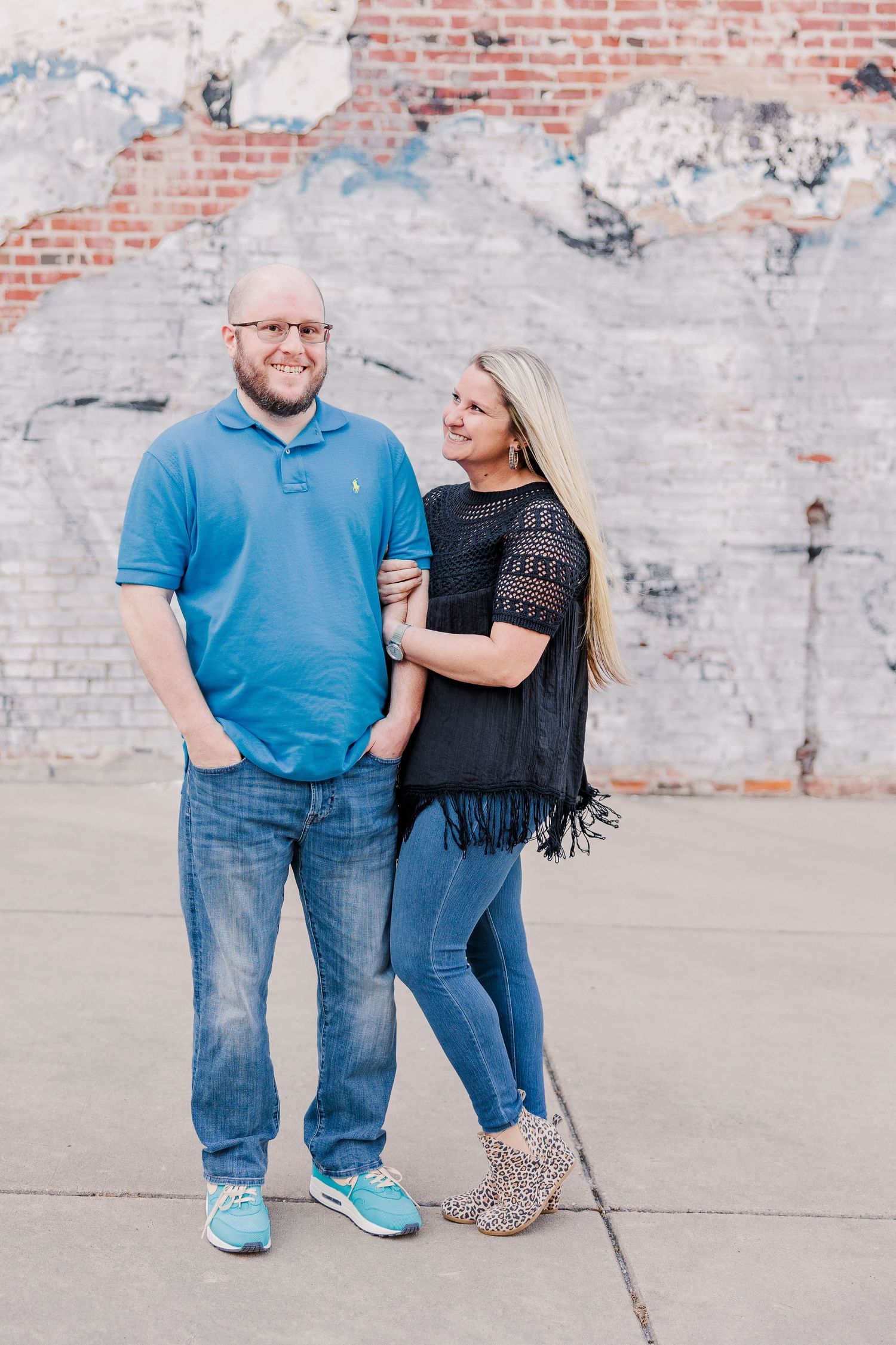 A couple poses together against a weathered brick wall, wearing casual blue attire.