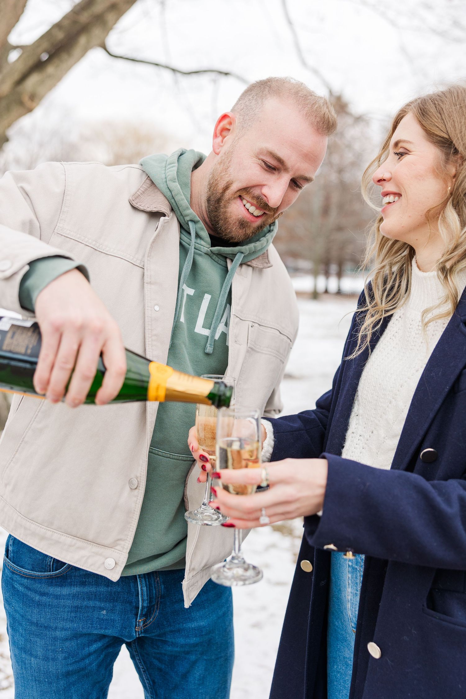 A couple shares a celebratory moment pouring champagne into glasses during a winter proposal photoshoot in Winnemac Park, Chicago.