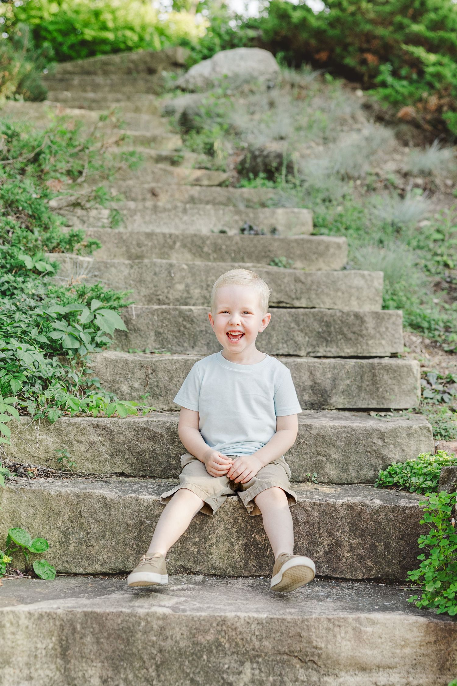 A small child wearing a gray t-shirt and shorts sits on stone garden steps surrounded by greenery.