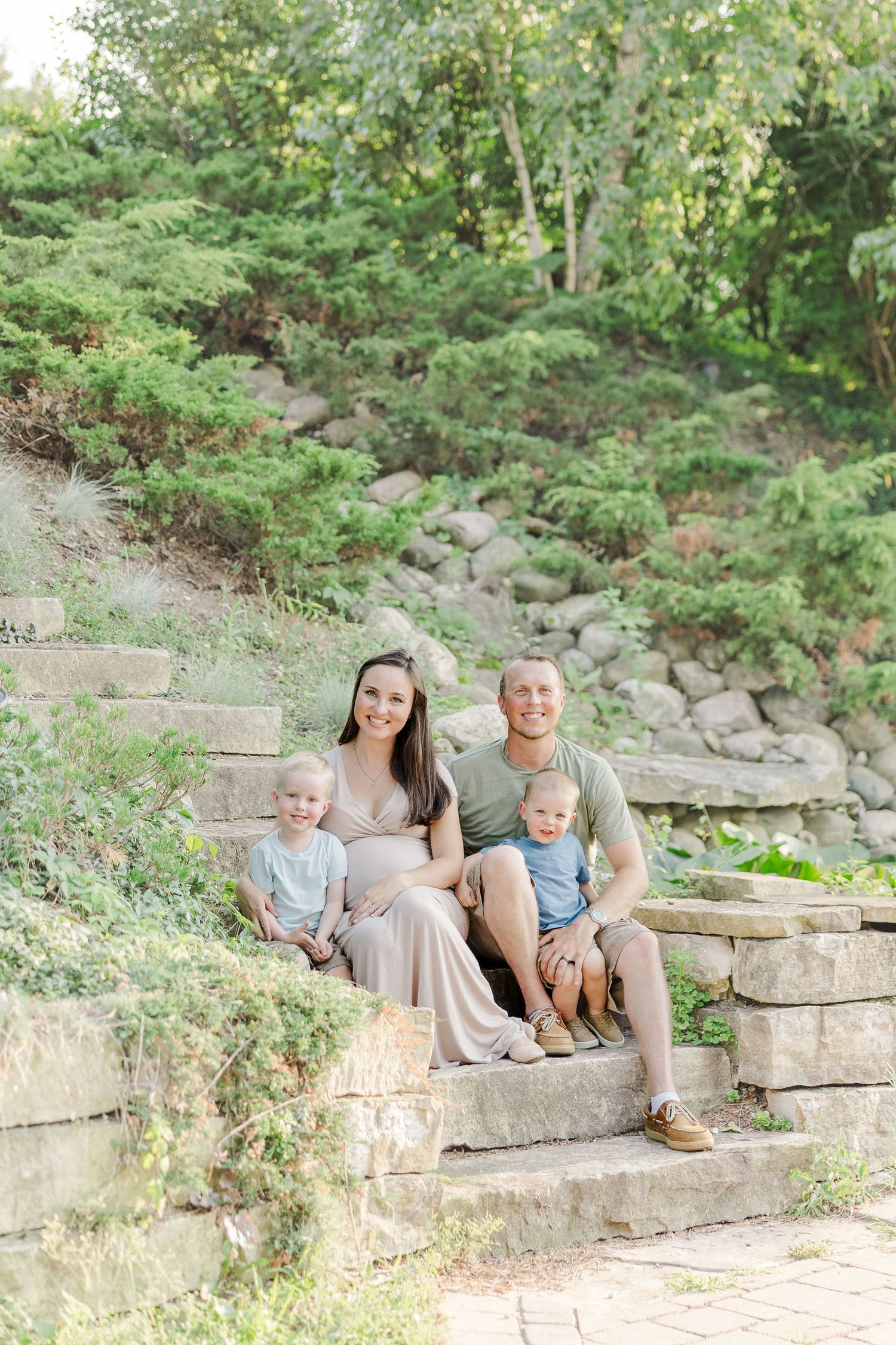 A young family sits together on stone garden steps surrounded by lush greenery on a bright summer day.
