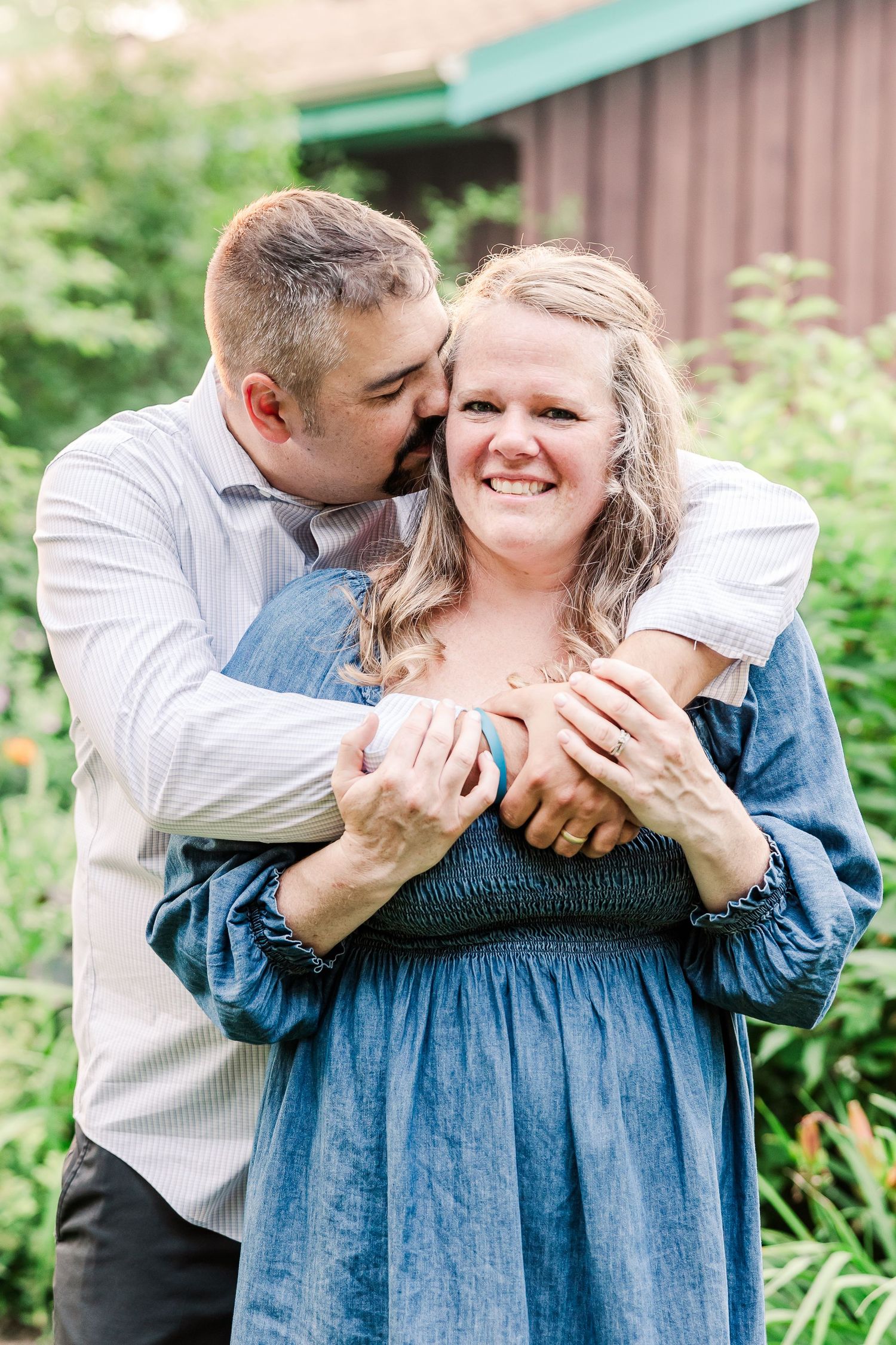 Romantic couple embrace in casual attire against garden backdrop.