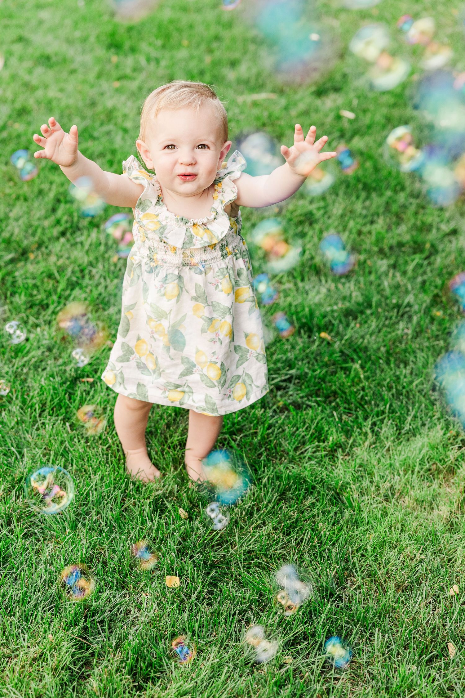 Toddler in floral dress reaches for bubbles floating in a sunny garden.
