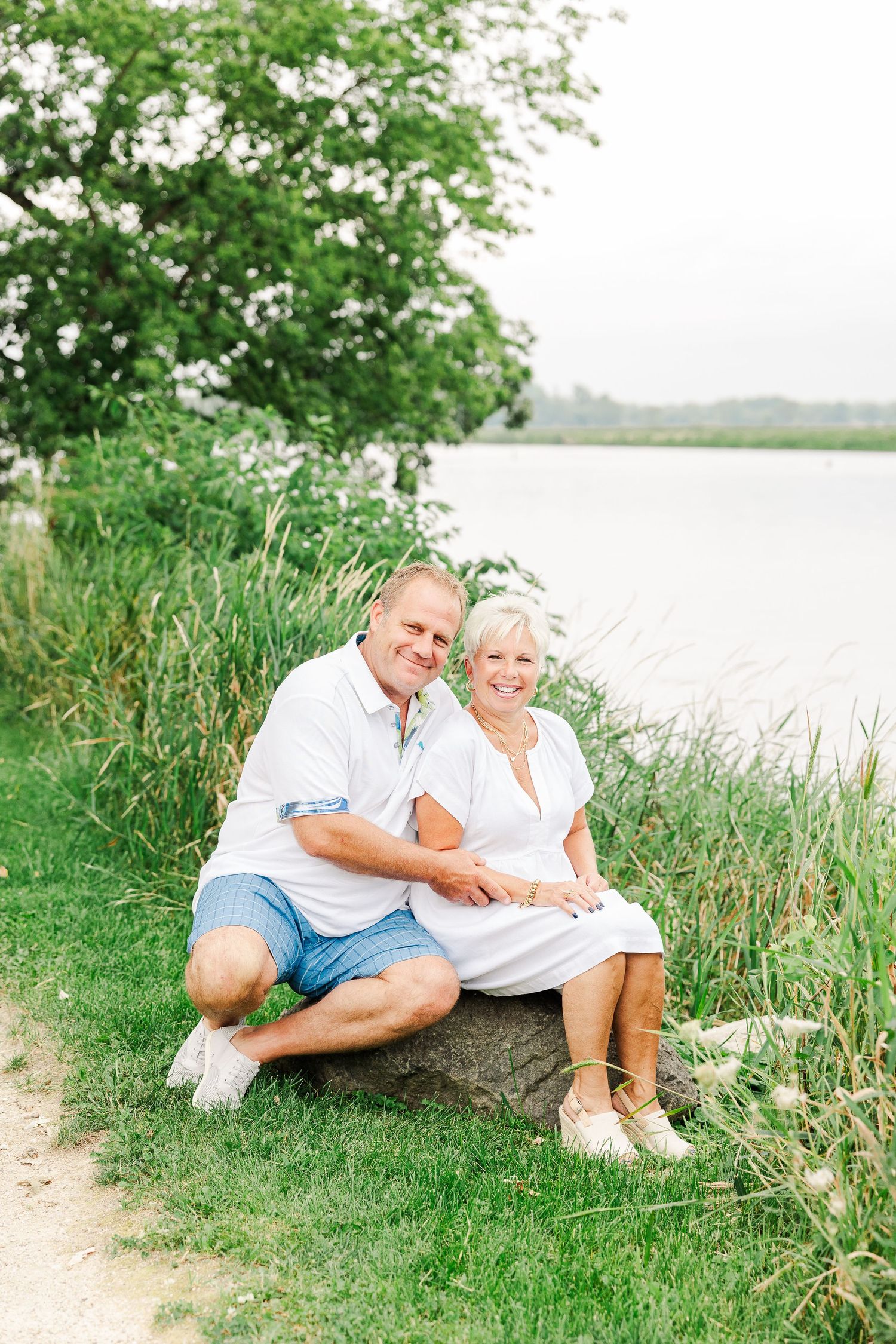 A couple relaxes on a rock by the waterfront surrounded by tall grass and trees.