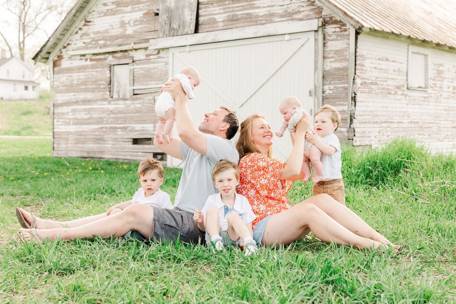 Playful family moment captured in front of a rustic wooden barn in a grassy field.