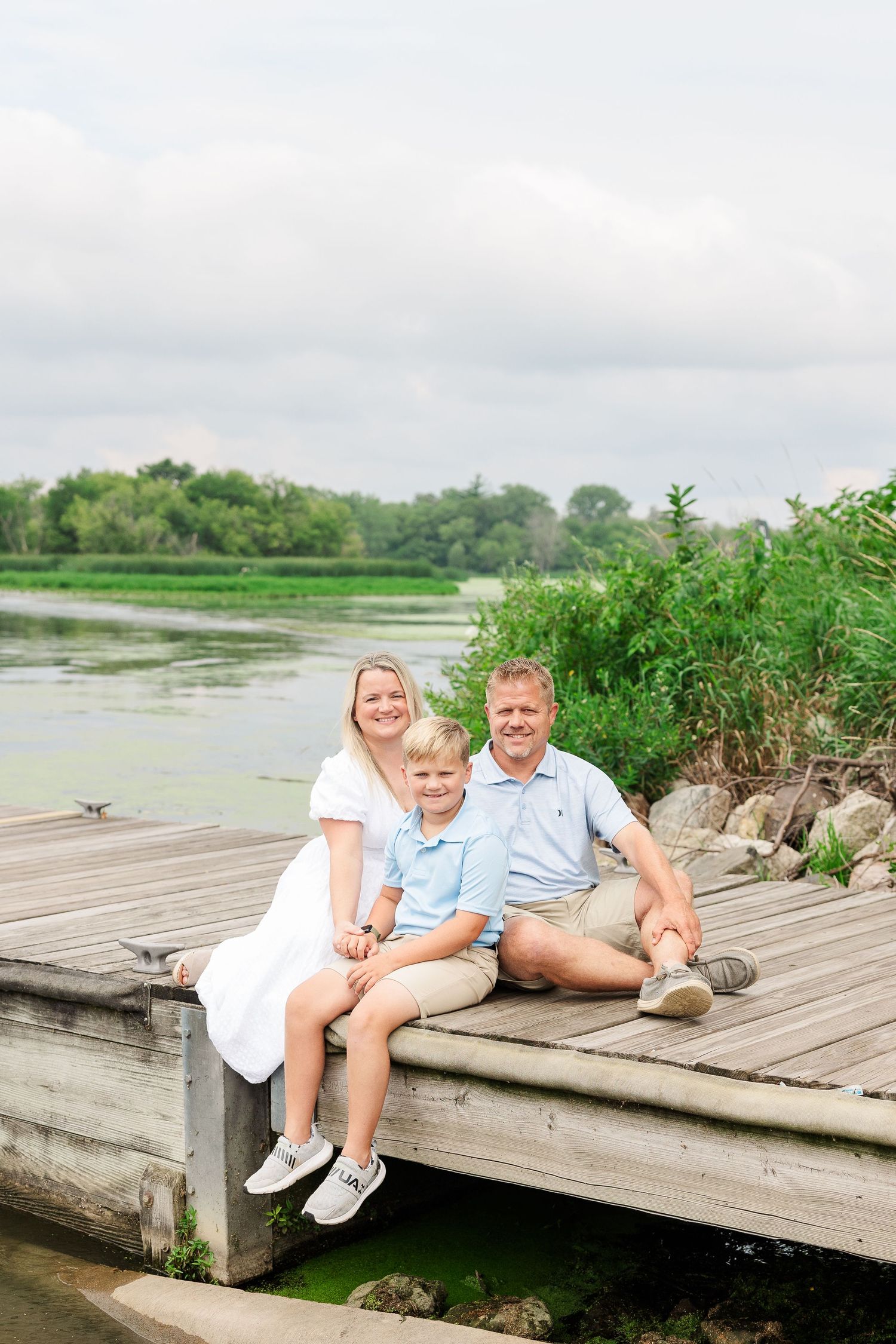 Family sitting casually on wooden dock edge overlooking peaceful lake and wooded shoreline.