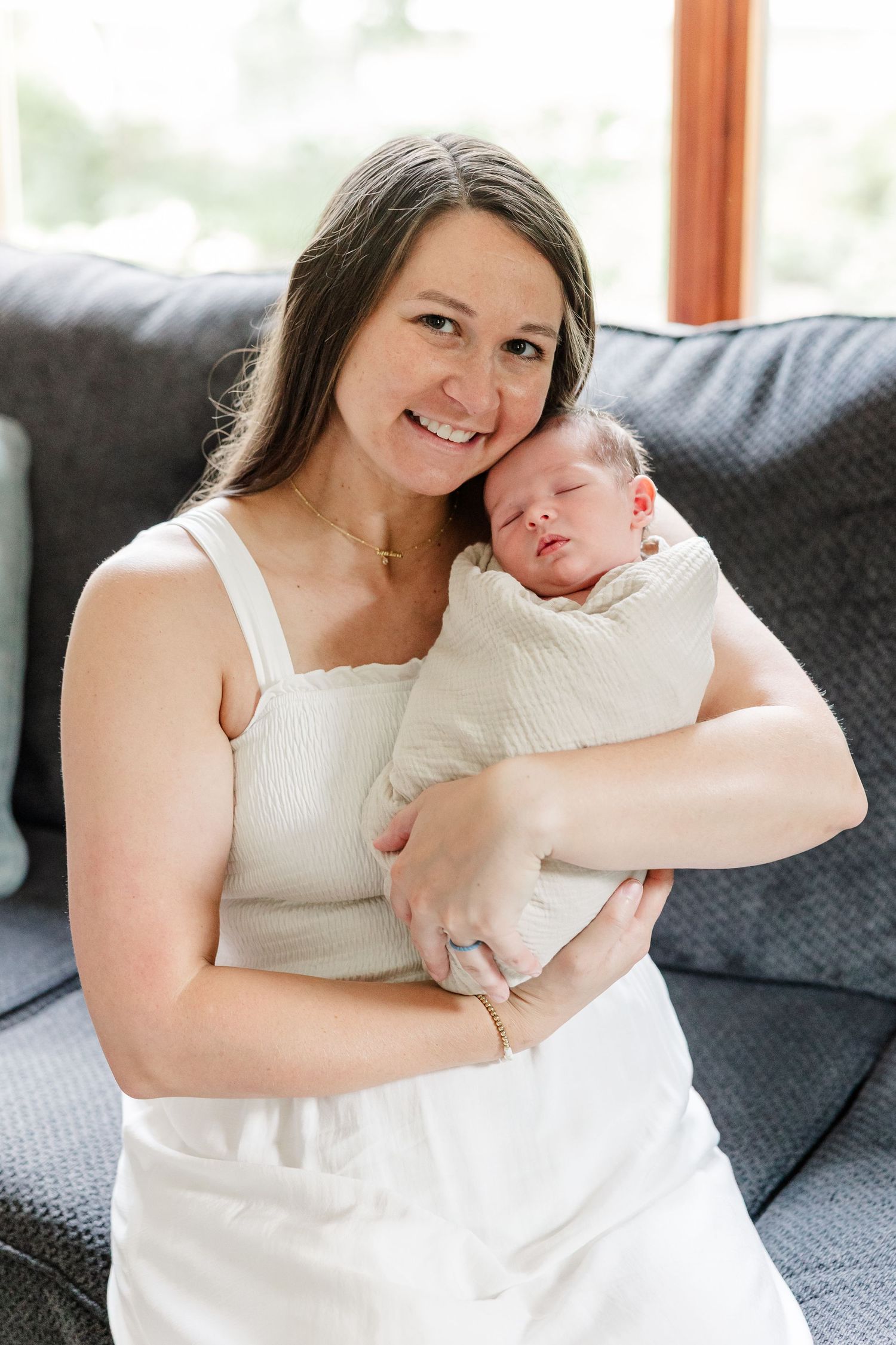 Smiling mother in white dress holds swaddled newborn baby while sitting on a gray couch indoors.
