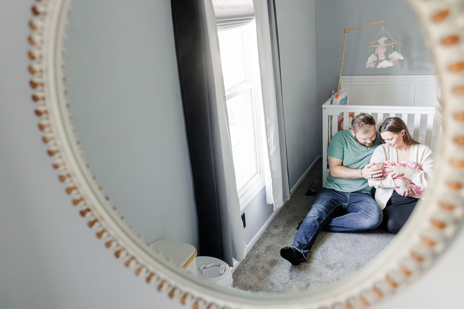 A mirror reflection shows new parents sitting on a nursery floor cradling their newborn baby near a white crib.