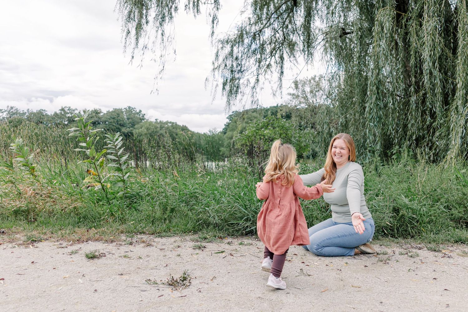 A smiling mother crouches down to hold hands with her young daughter near a weeping willow tree outdoors.