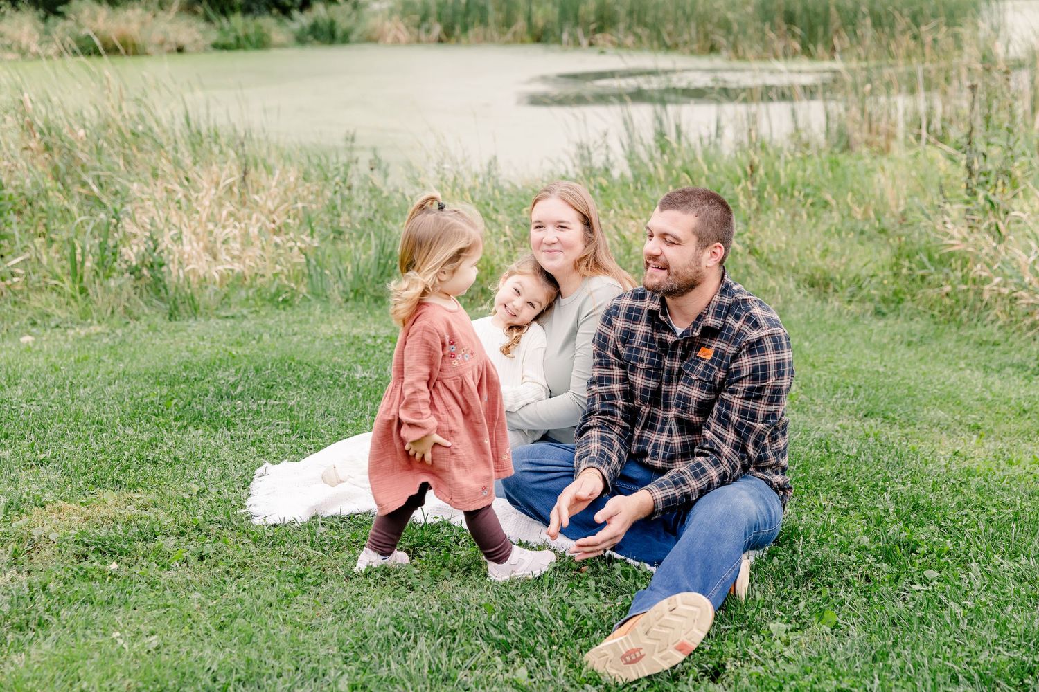 Family of four sitting on grass by a pond, smiling and interacting outdoors on a warm autumn day.