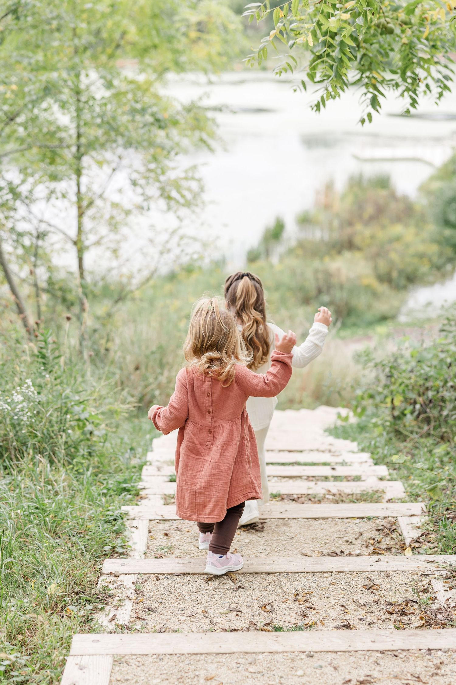 Two young girls walking up outdoor stone steps surrounded by lush greenery near a lake.