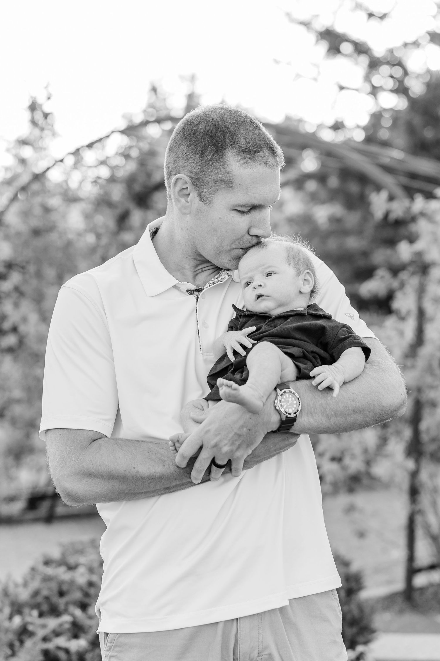 Black-and-white portrait of Andrew kissing newborn Lincoln while holding him outdoors.