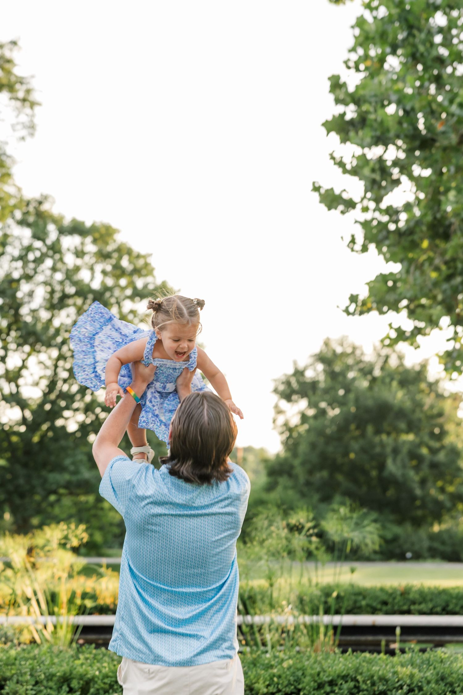 Father lifts laughing toddler daughter high in air against bright sky in lush green park setting.