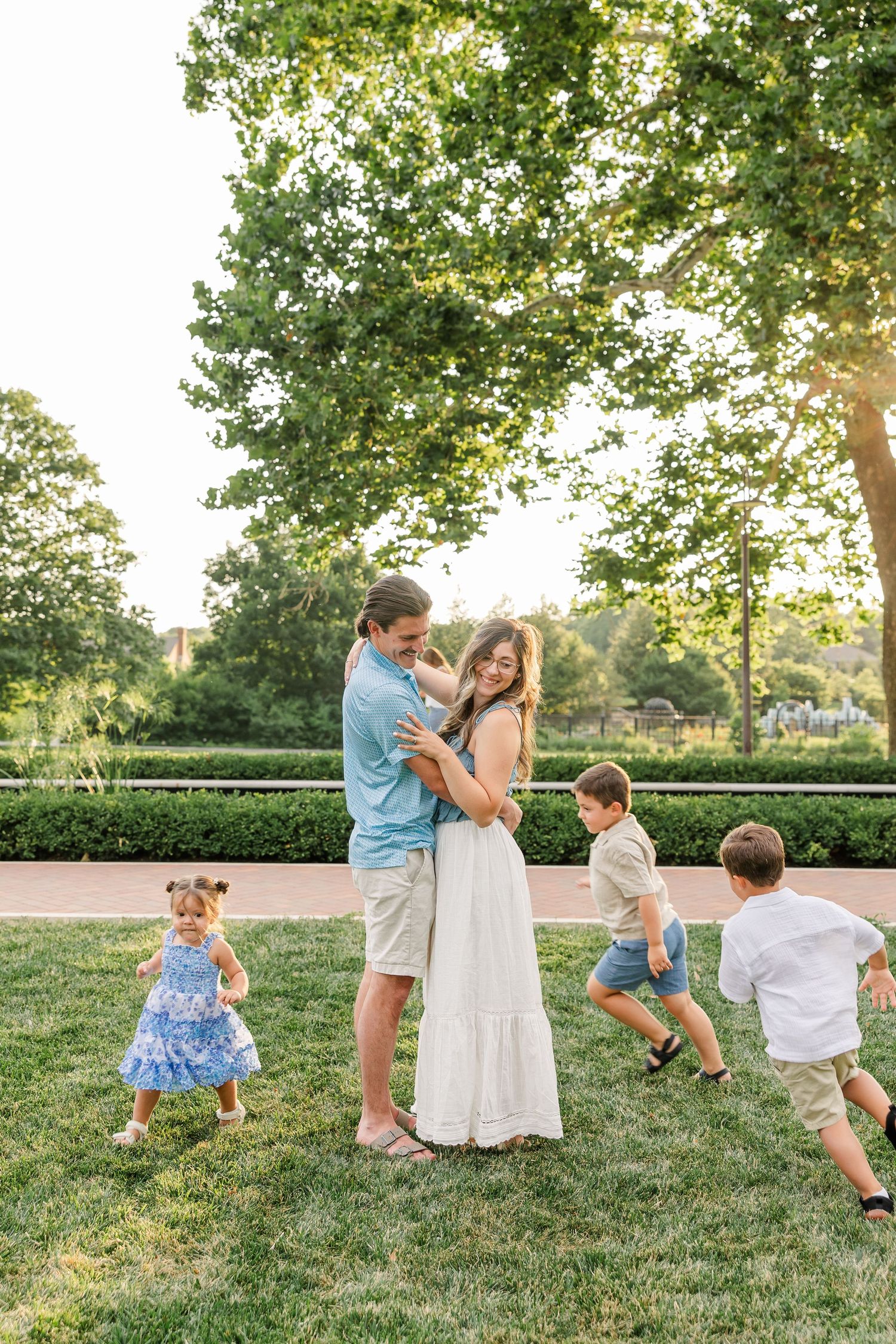 A happy couple embraces on green grass while their three young children play around them in golden light.