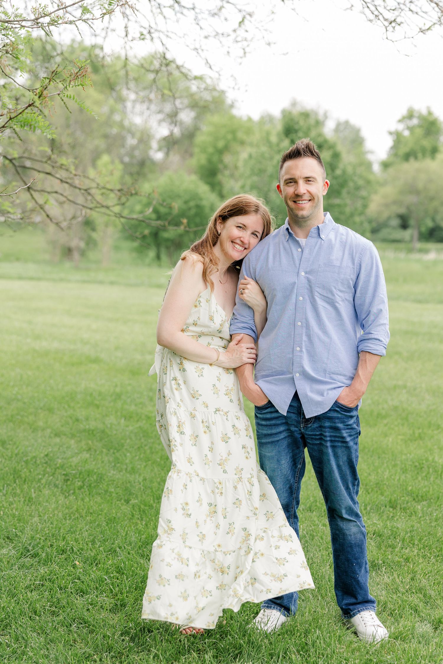 Smiling couple in casual attire standing together on green grass under spring tree branches outdoors.