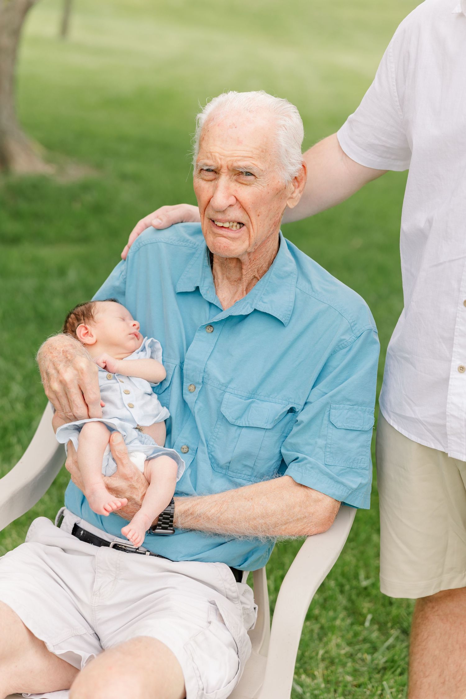 Elderly grandfather in blue shirt holds sleeping newborn baby while seated in white chair outdoors.